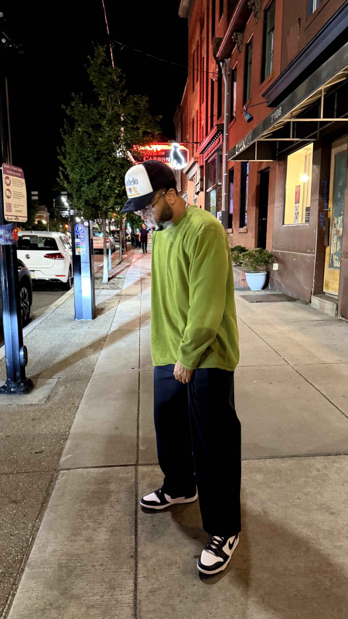 Person standing on sidewalk at night, wearing a green sweater, black pants, and sneakers, with buildings and streetlights in background.