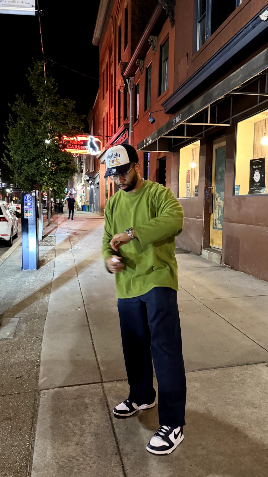 Man in green sweater, dark pants, and sneakers standing on city sidewalk at night, looking at his watch.