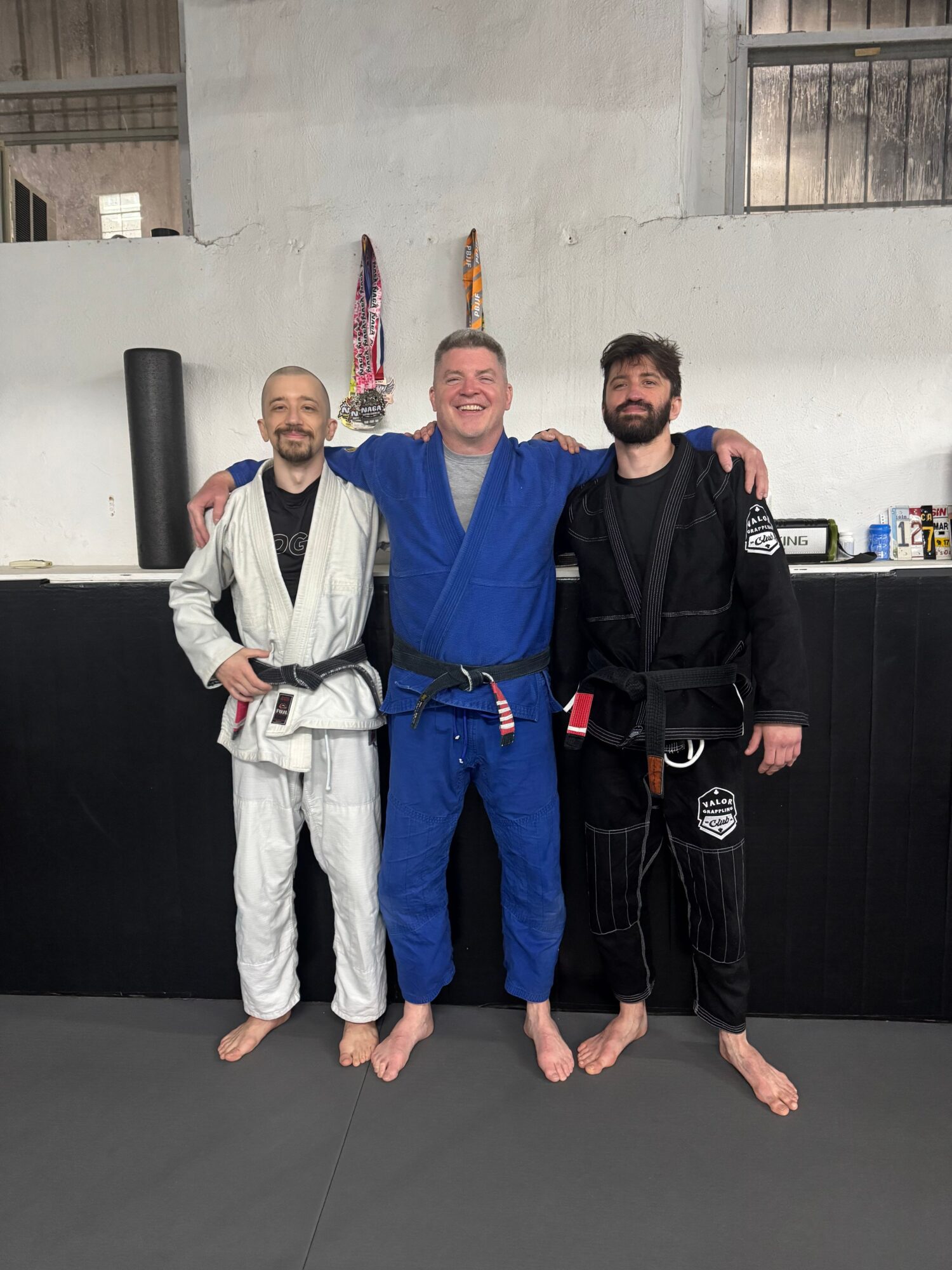 Three people in martial arts uniforms and a person in a black jacket standing on a gray mat, with a concrete wall in the background.