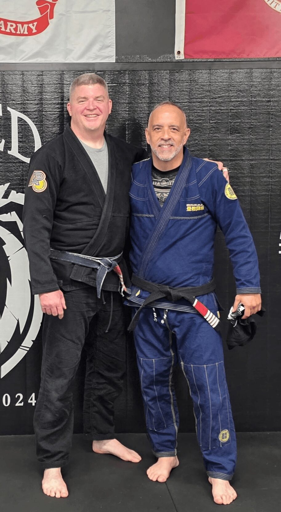 Two men in martial arts uniforms standing side by side, smiling, with black mats and banners in the background.