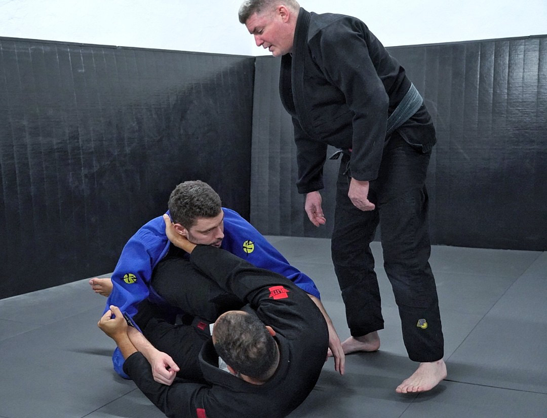Two people practicing Brazilian Jiu-Jitsu on a mat, one in a blue gi and the other in a black gi, with a third person standing nearby. Short-haired man in black gi is observing.