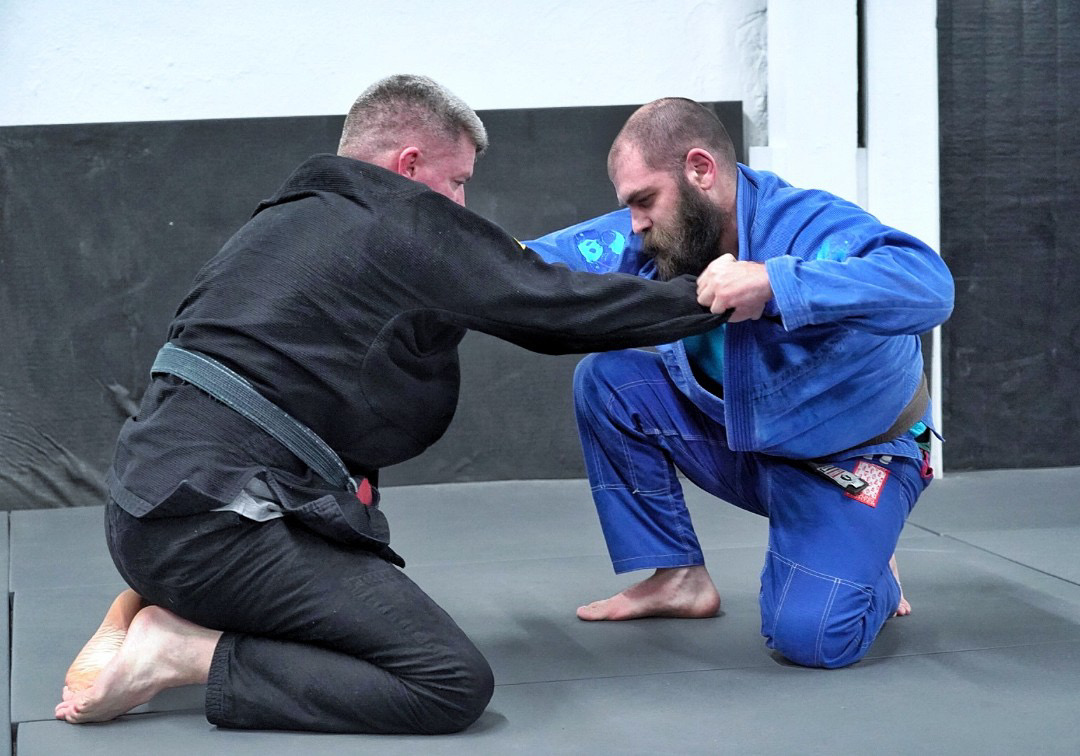 Two men practicing Brazilian Jiu-Jitsu on a mat, one in a black gi and the other in a blue gi, engaged in a grappling position.