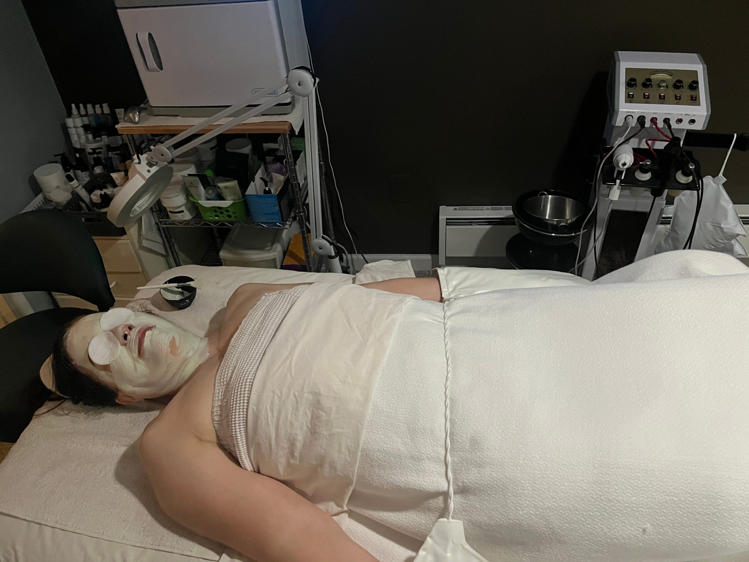 Person lying in a medical or spa setting with face mask, wrapped in a towel, surrounded by equipment and supplies.