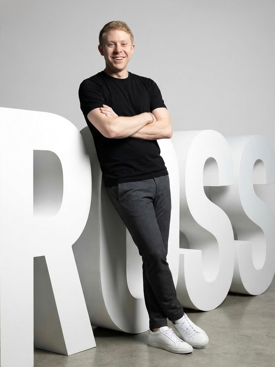 Young man with crossed arms leaning against large white letters, smiling, in a gray studio background.