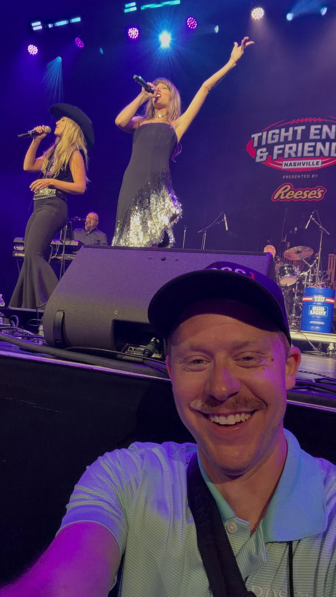 Performer singing on stage with a microphone, another person in the background, and a smiling man in the foreground wearing a cap.