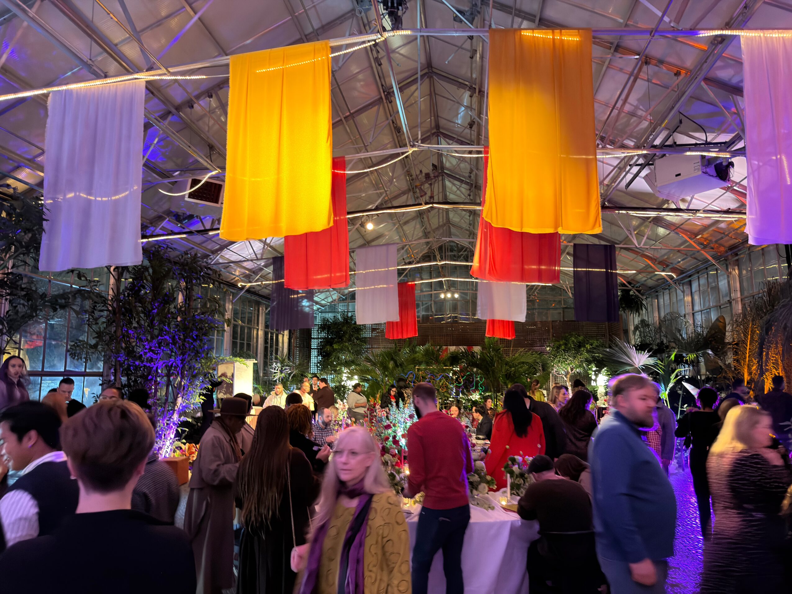 Indoor event with hanging yellow, red, and purple fabric decorations, and people socializing around tables.