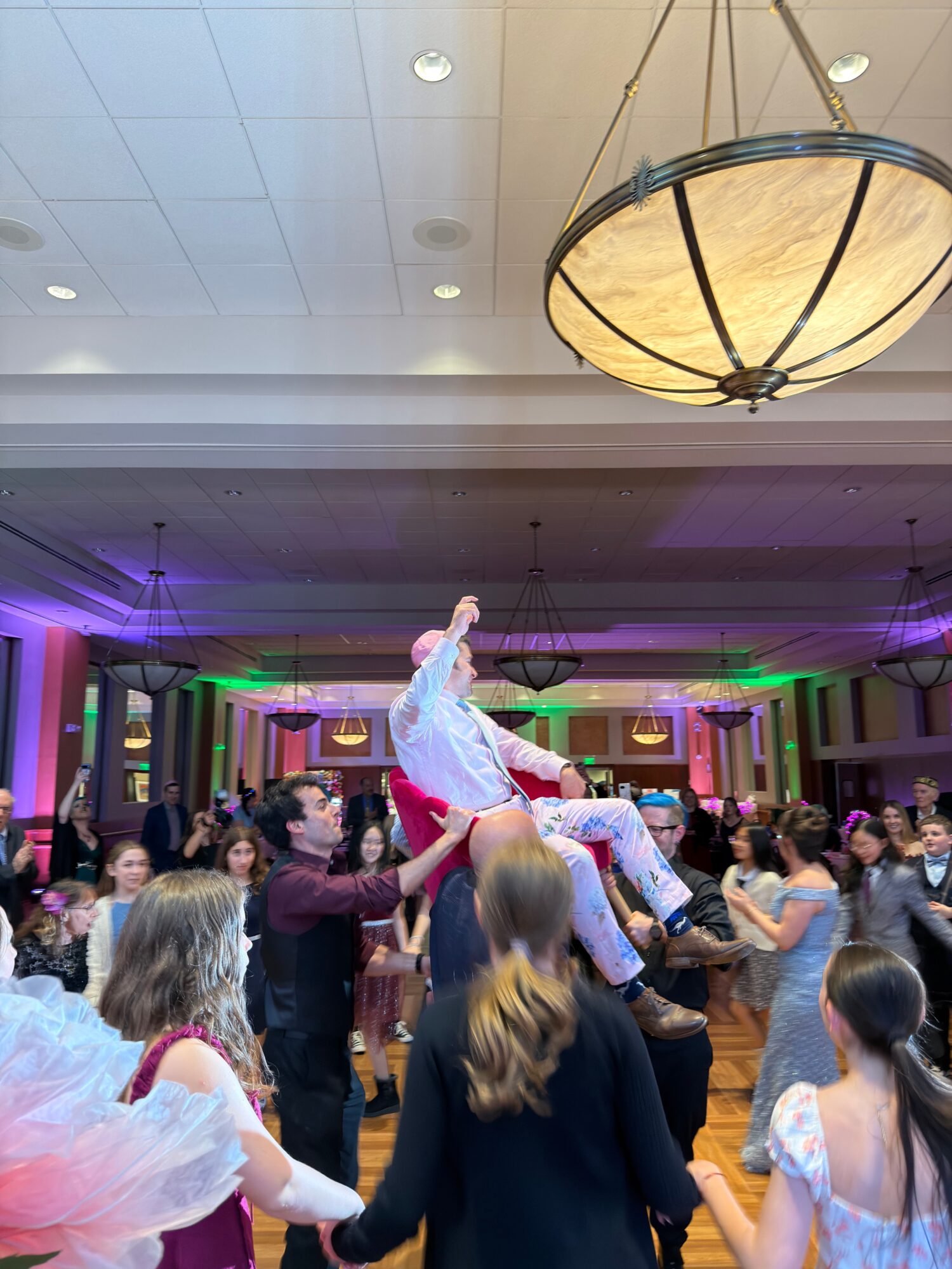 People dancing and socializing in a decorated indoor venue with colorful lighting and a high ceiling.
