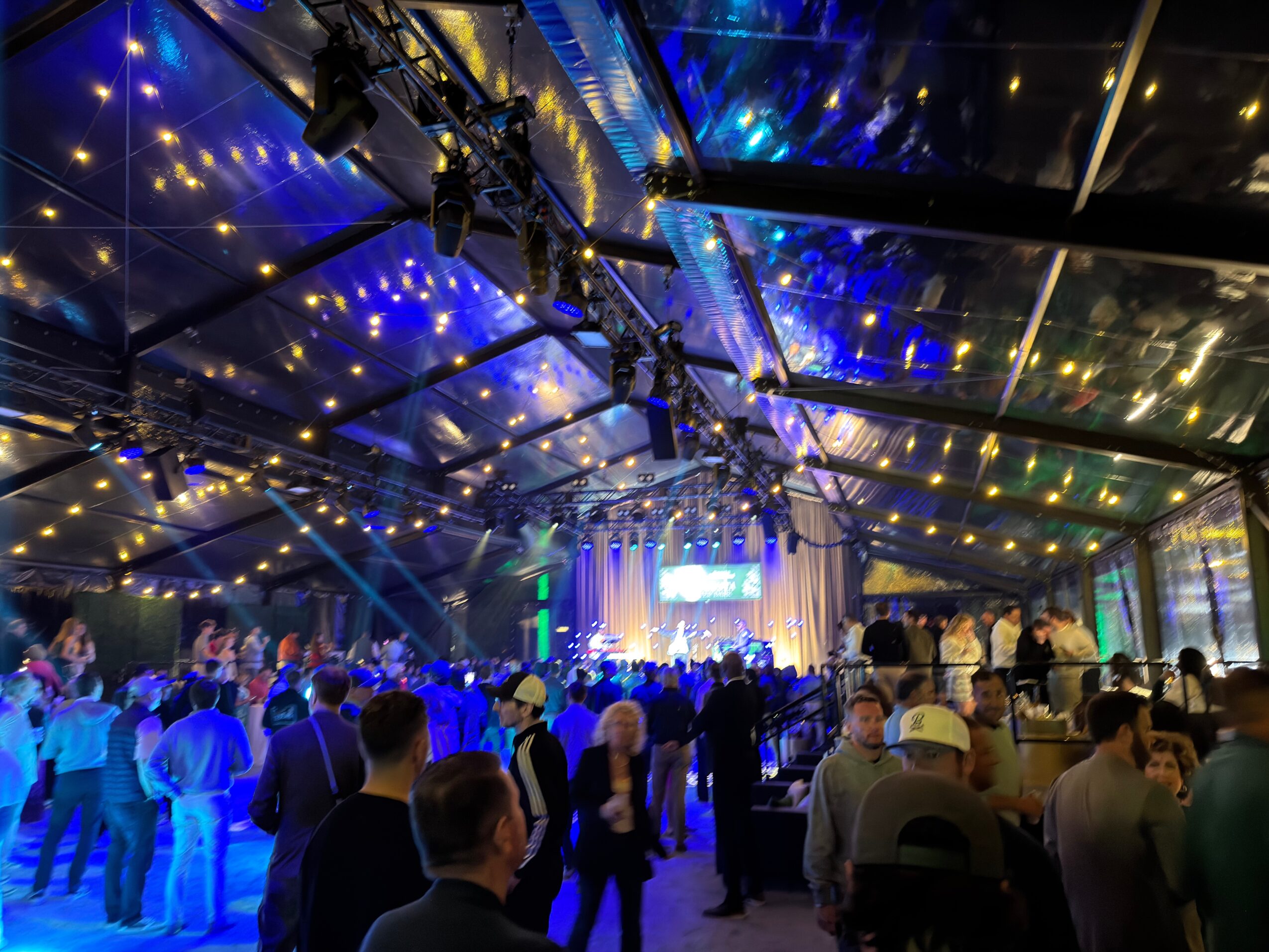 Crowd of people inside a large, decorated event tent with blue and yellow lighting, stage at the front, and ceiling lights.