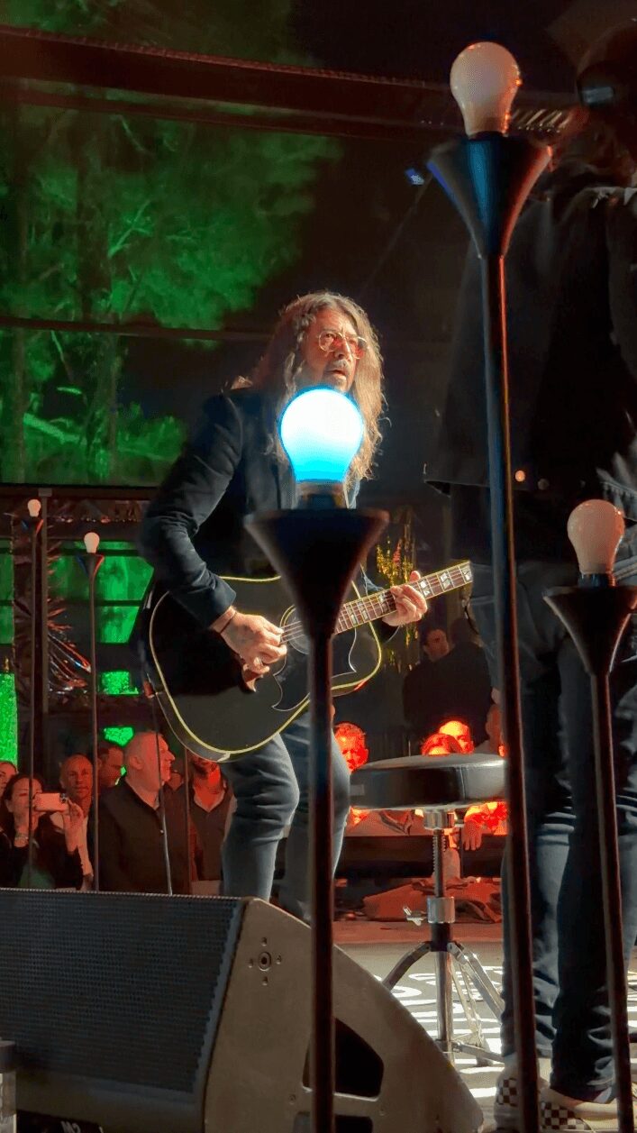 Musician with long hair playing guitar on stage with colorful lighting and audience in background.