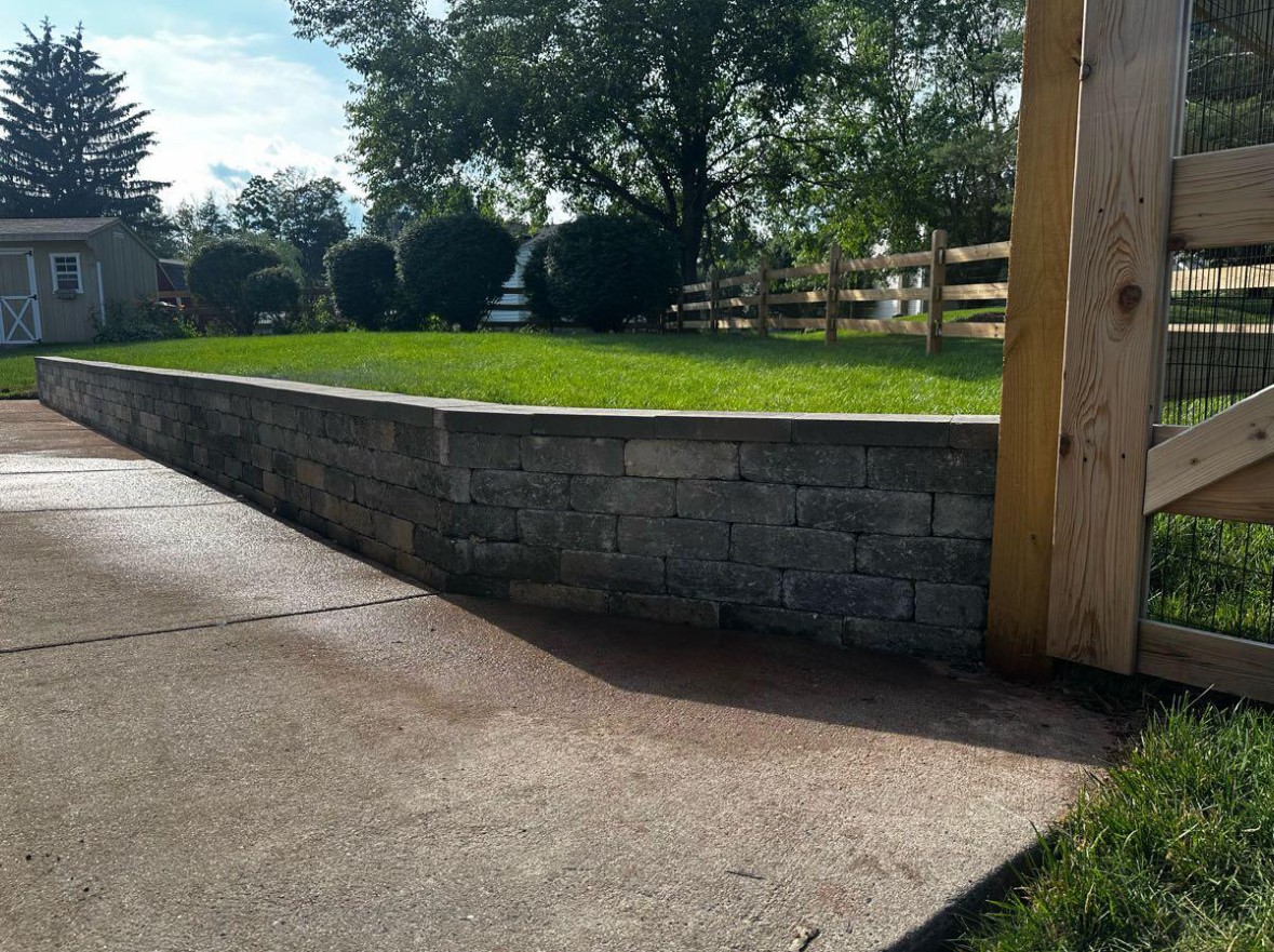 Stone retaining wall with grass on top, adjacent to a concrete sidewalk and wooden fence, in a sunny outdoor setting.