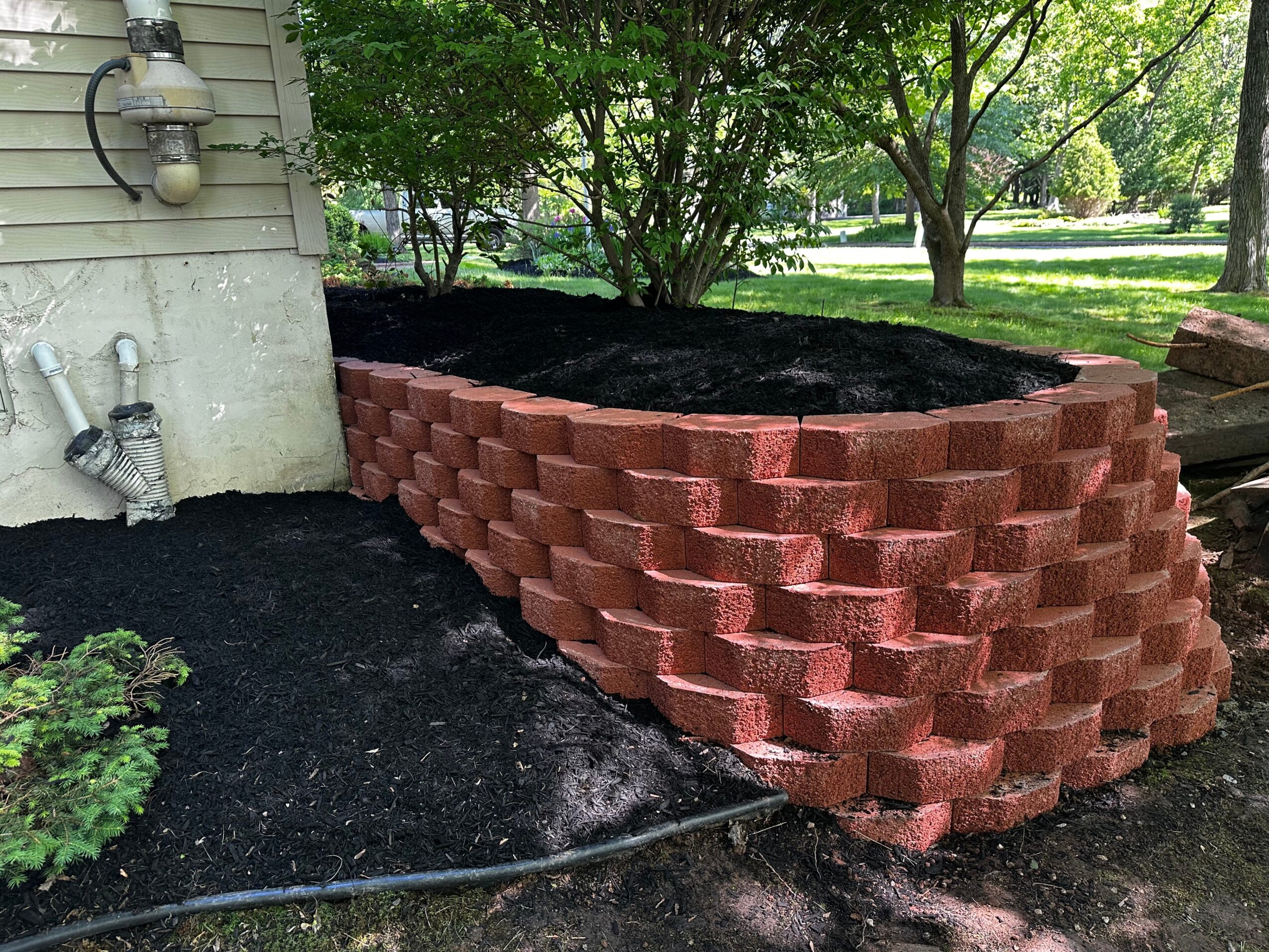 Red brick garden bed with soil, next to house wall and trees in background.