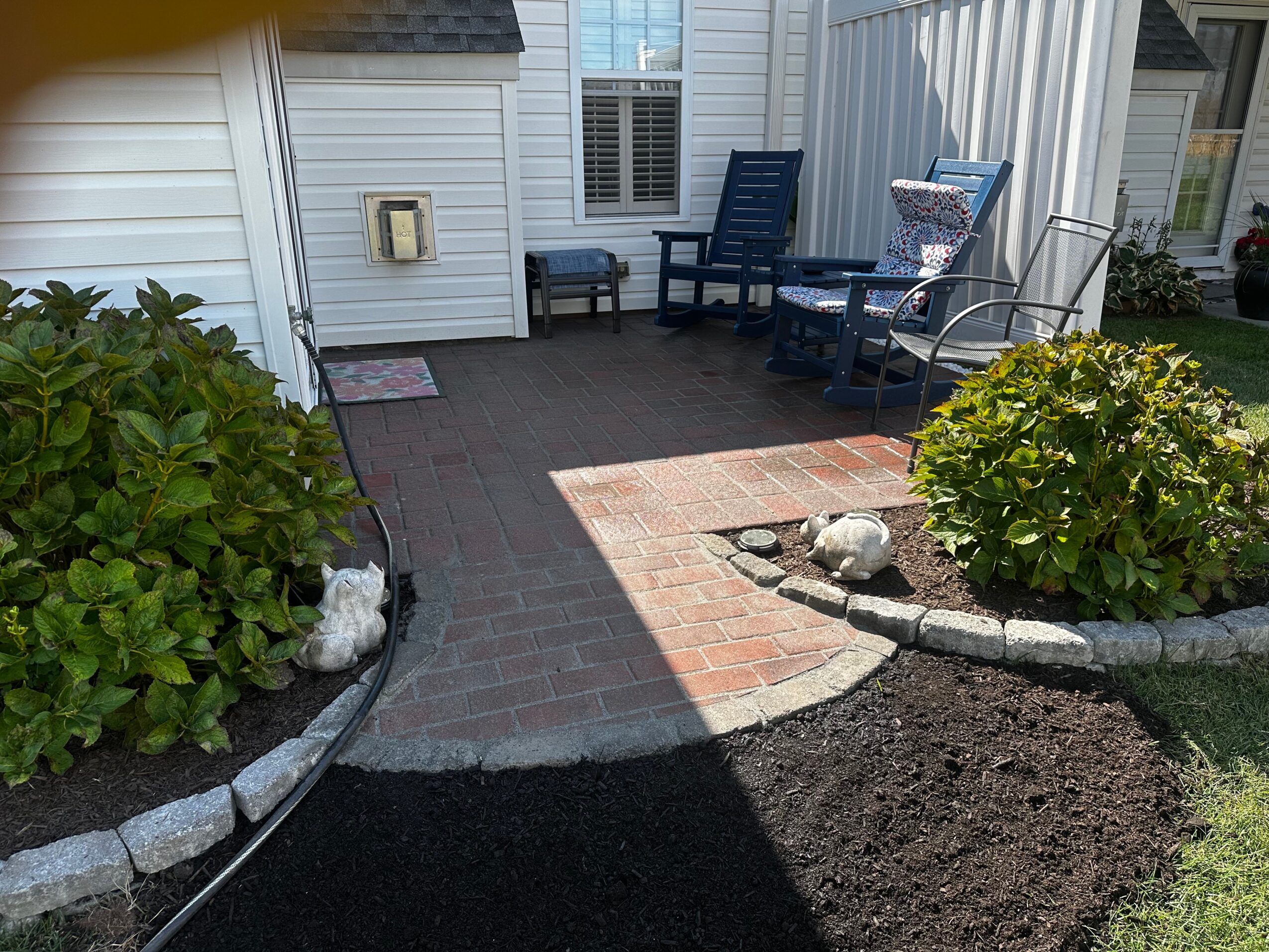 Patio with chairs, bushes, and a brick pathway outside a house, sunlight casting shadows, and a garden bed with rocks.