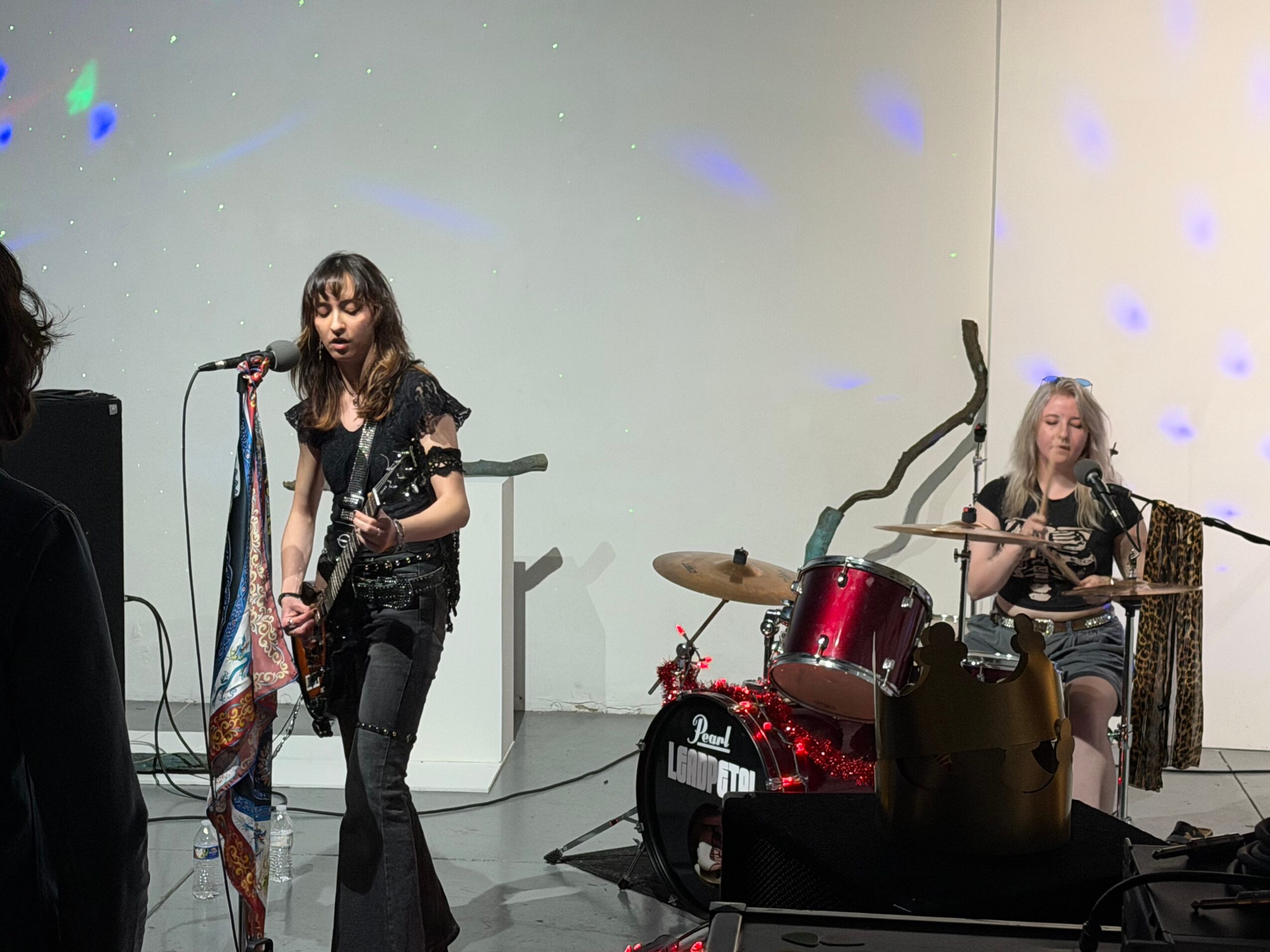 Two young girls perform on stage with musical instruments, one singing and playing guitar, the other on drums, in a simple setting.