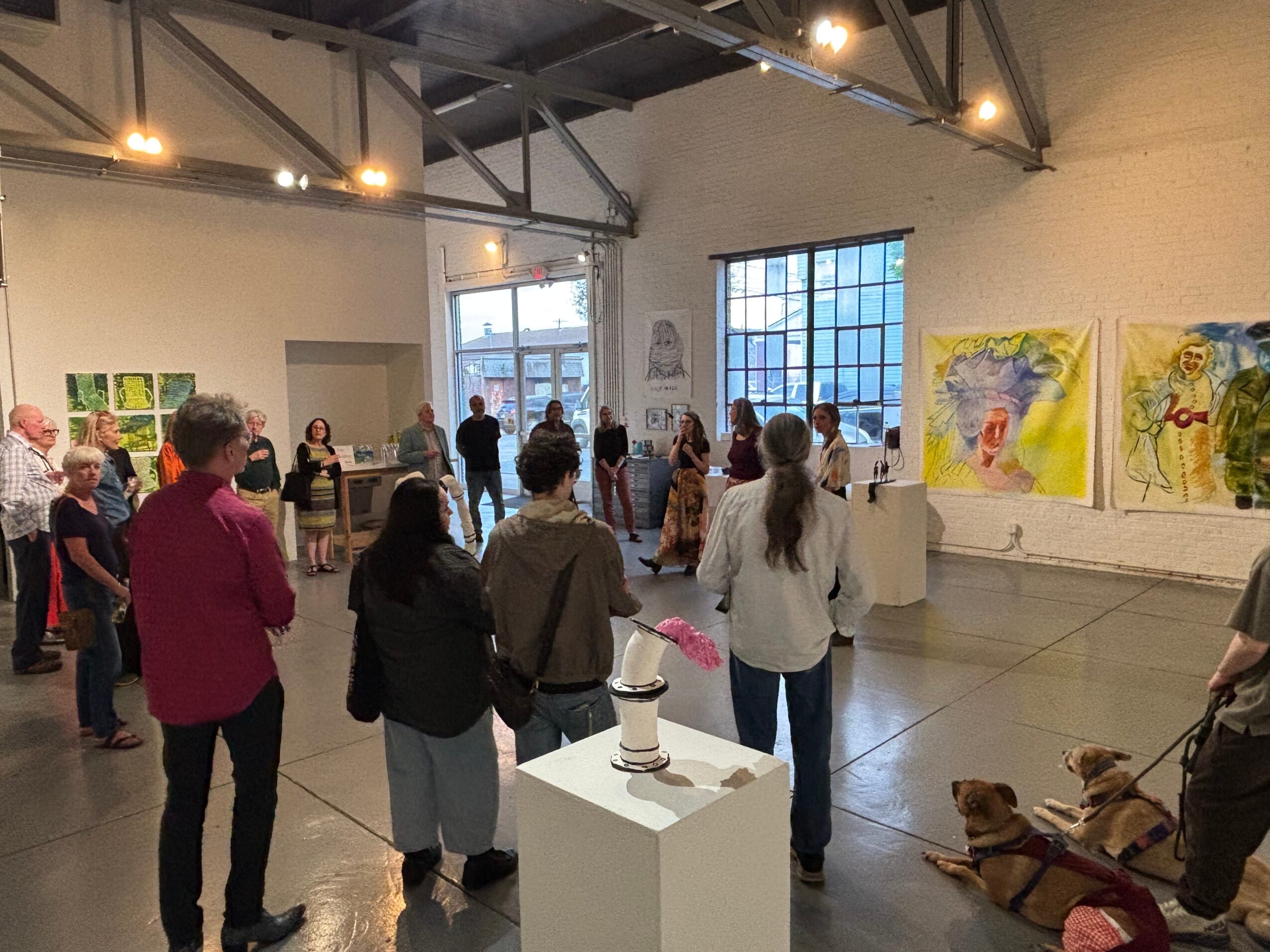 People gathered in an art gallery viewing paintings and sculptures, with large windows and high ceilings.