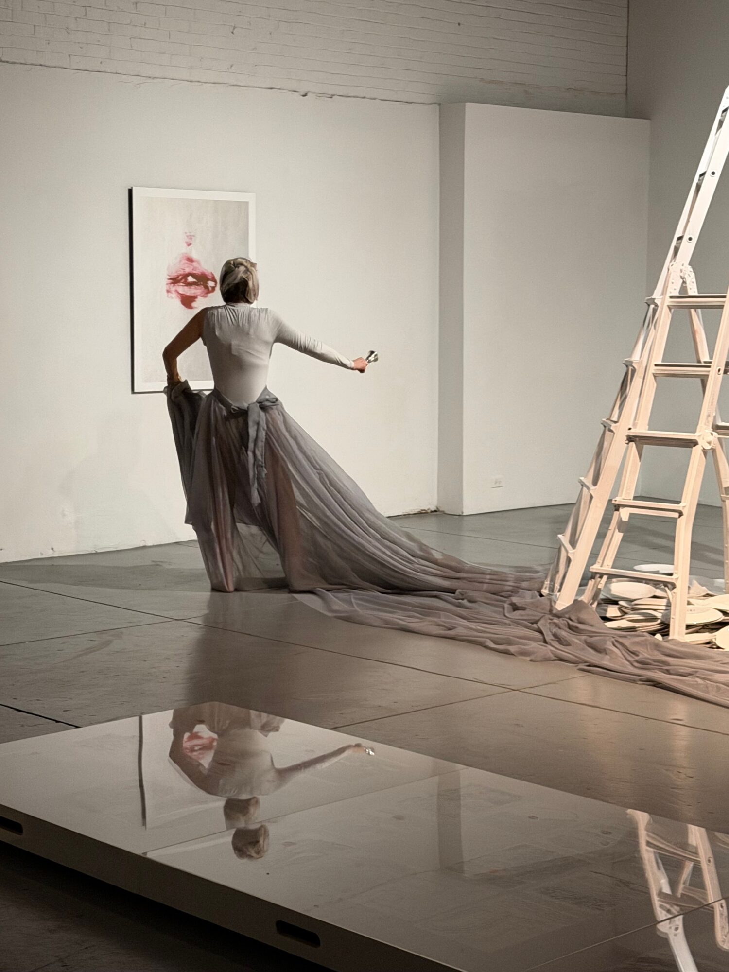 Person standing in an art gallery, looking at artwork on the wall, with their reflection visible in a mirror.