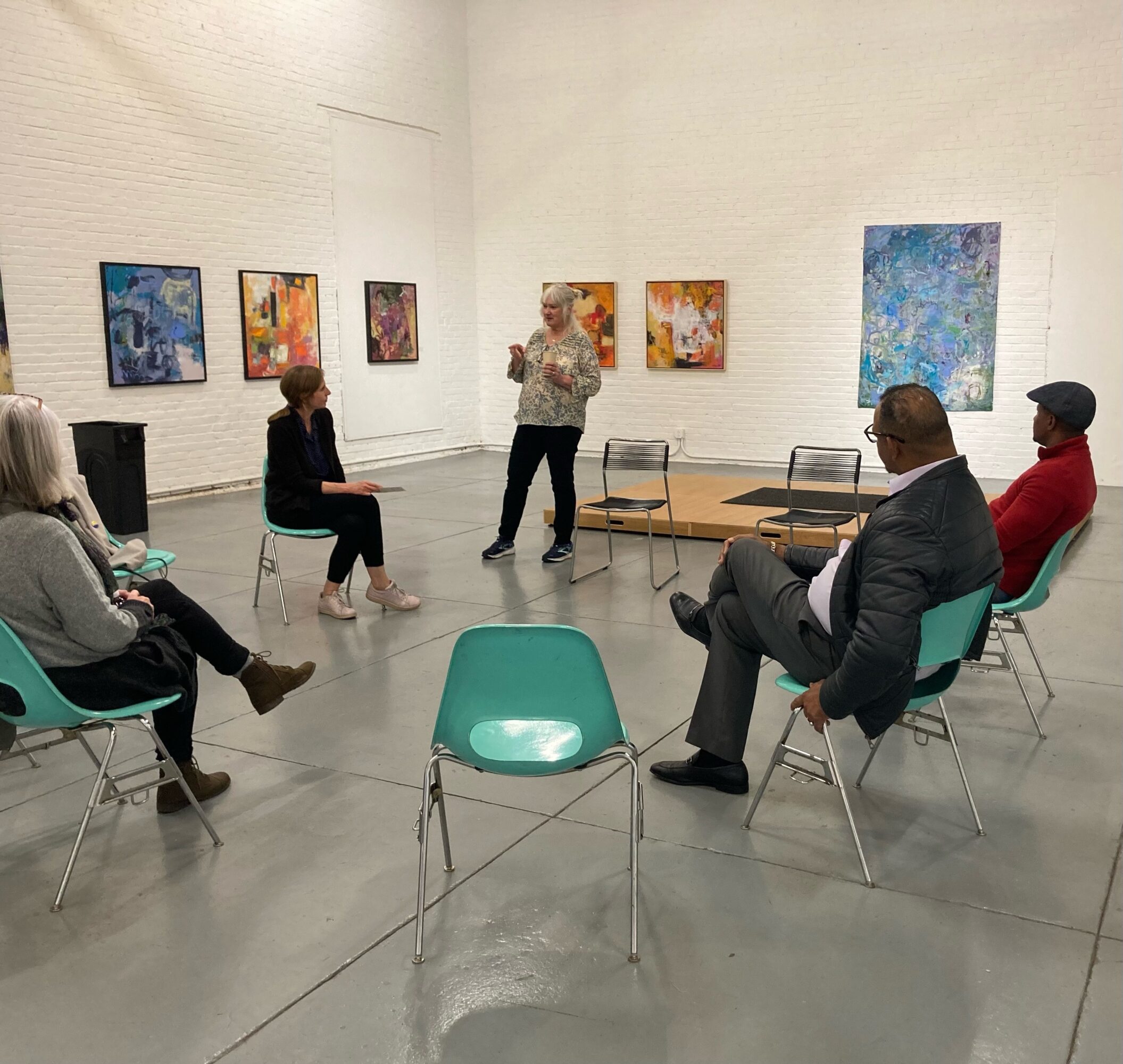 Group of six people seated in a circle in an art gallery, listening to a woman speaking, colorful paintings on white walls.