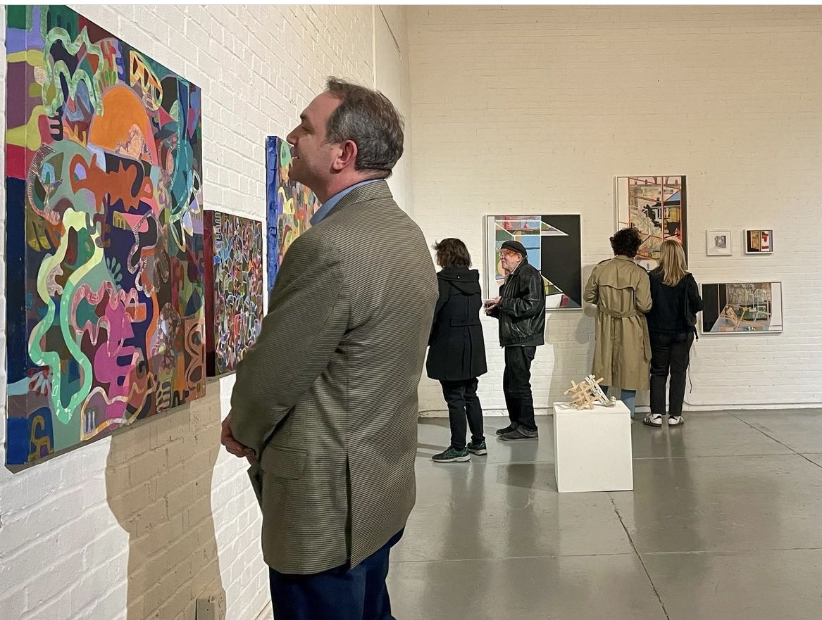 Man in gray blazer looking at colorful abstract paintings in art gallery with other visitors in background.