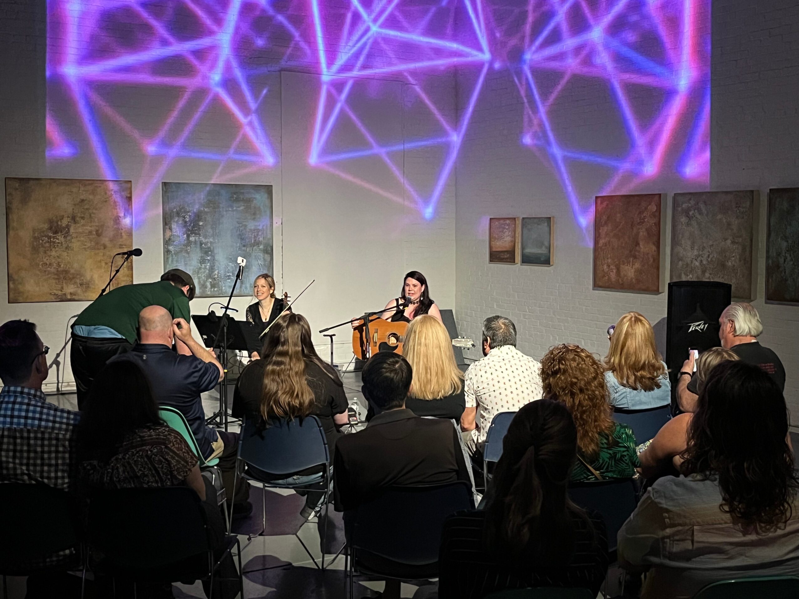 Three musicians perform on stage with audience watching, colorful abstract art on walls, purple geometric light projection above, in an indoor venue.