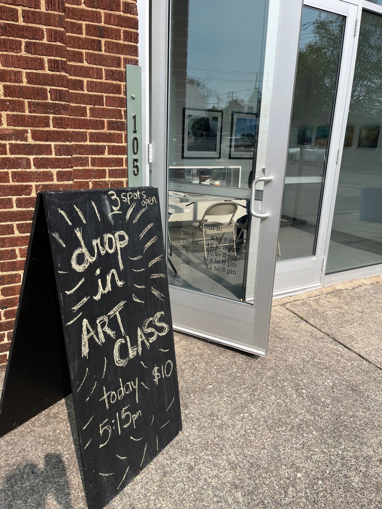 Black sidewalk sign with white handwritten text outside a building, brick wall and glass door visible, shadow on ground.