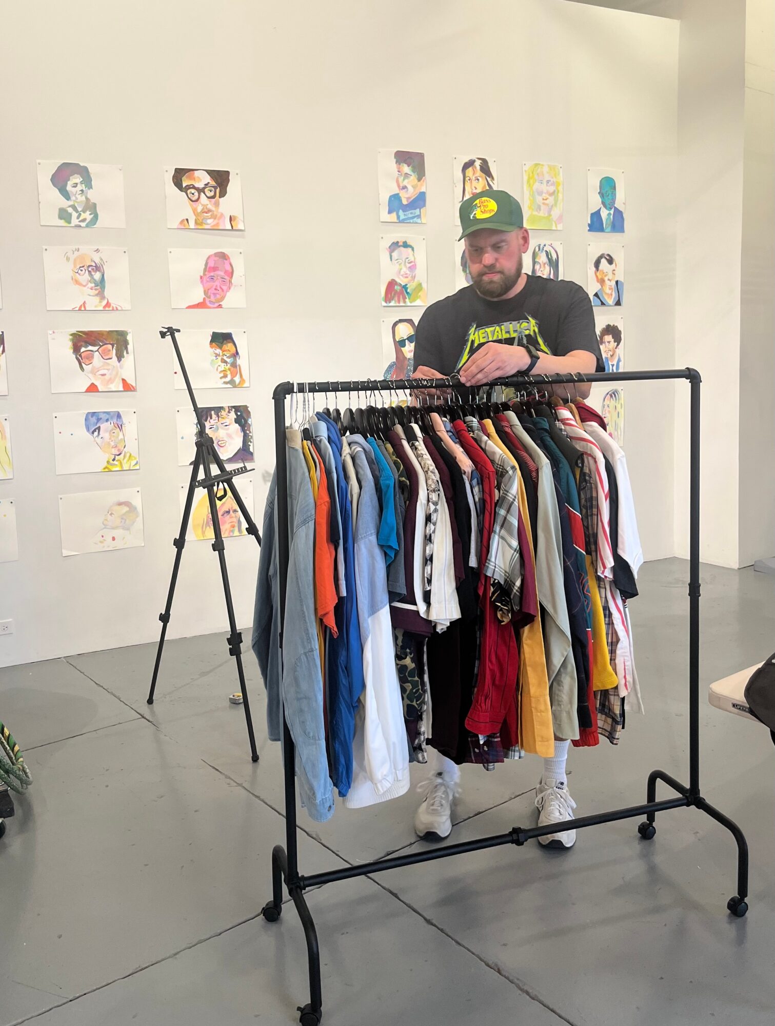Person with a beard and cap stands behind a clothing rack filled with various shirts, in an art gallery with colorful portraits on the wall.