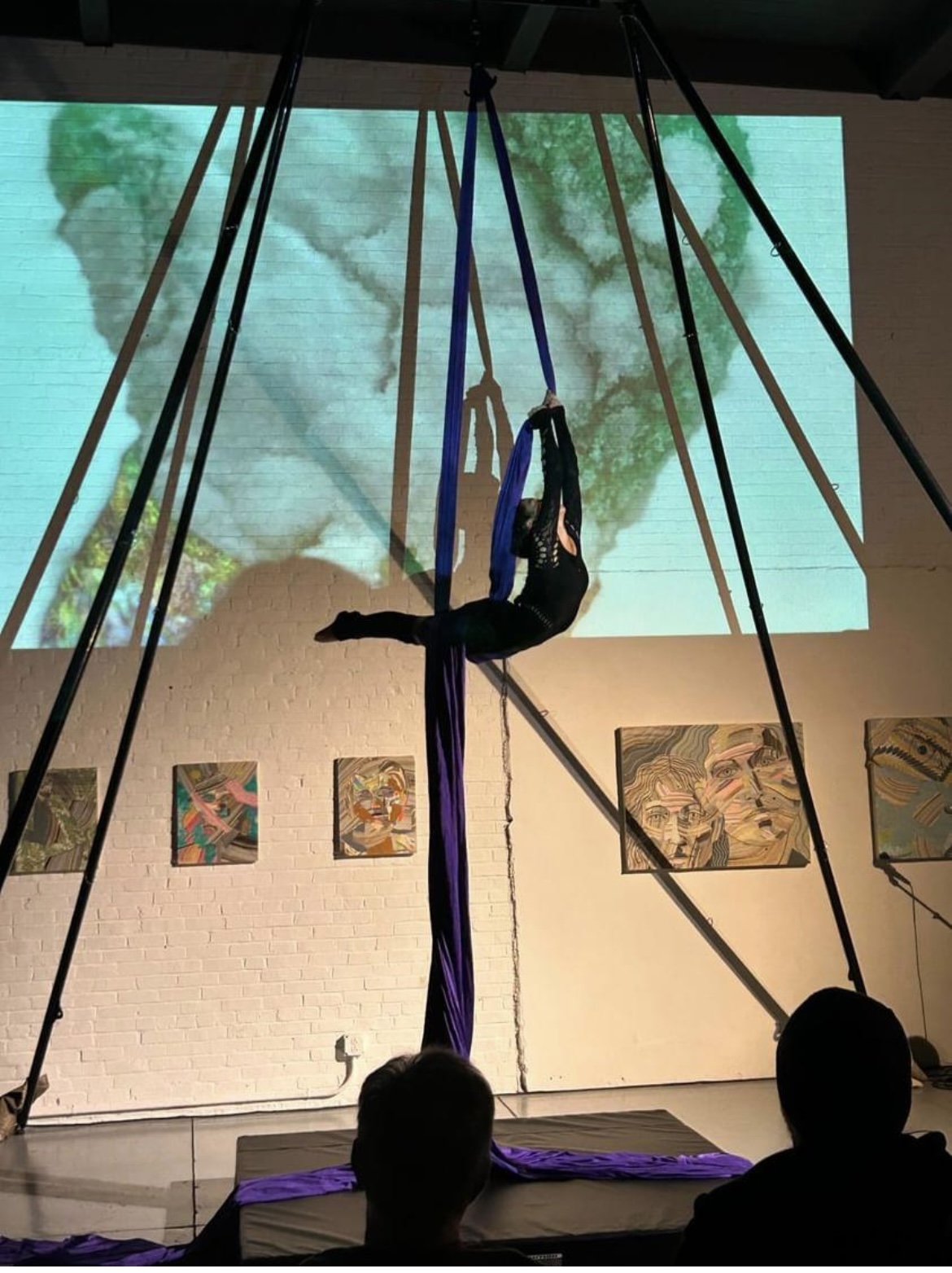 Aerial performer hangs upside down on fabric, with a large screen showing a cloud scene behind, in an indoor venue.