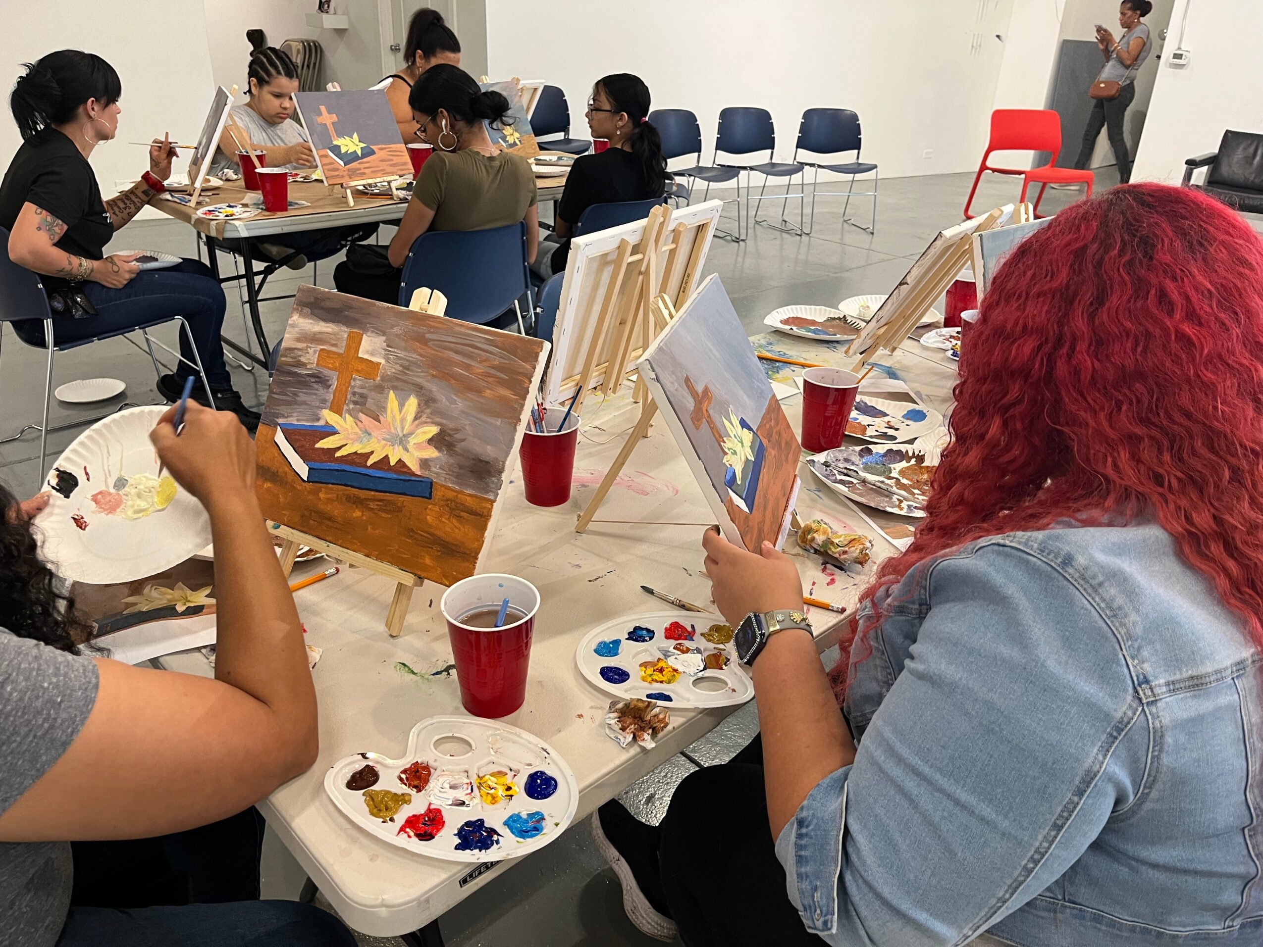 People painting on canvases at a table with paint palettes and drinks, in a bright room.