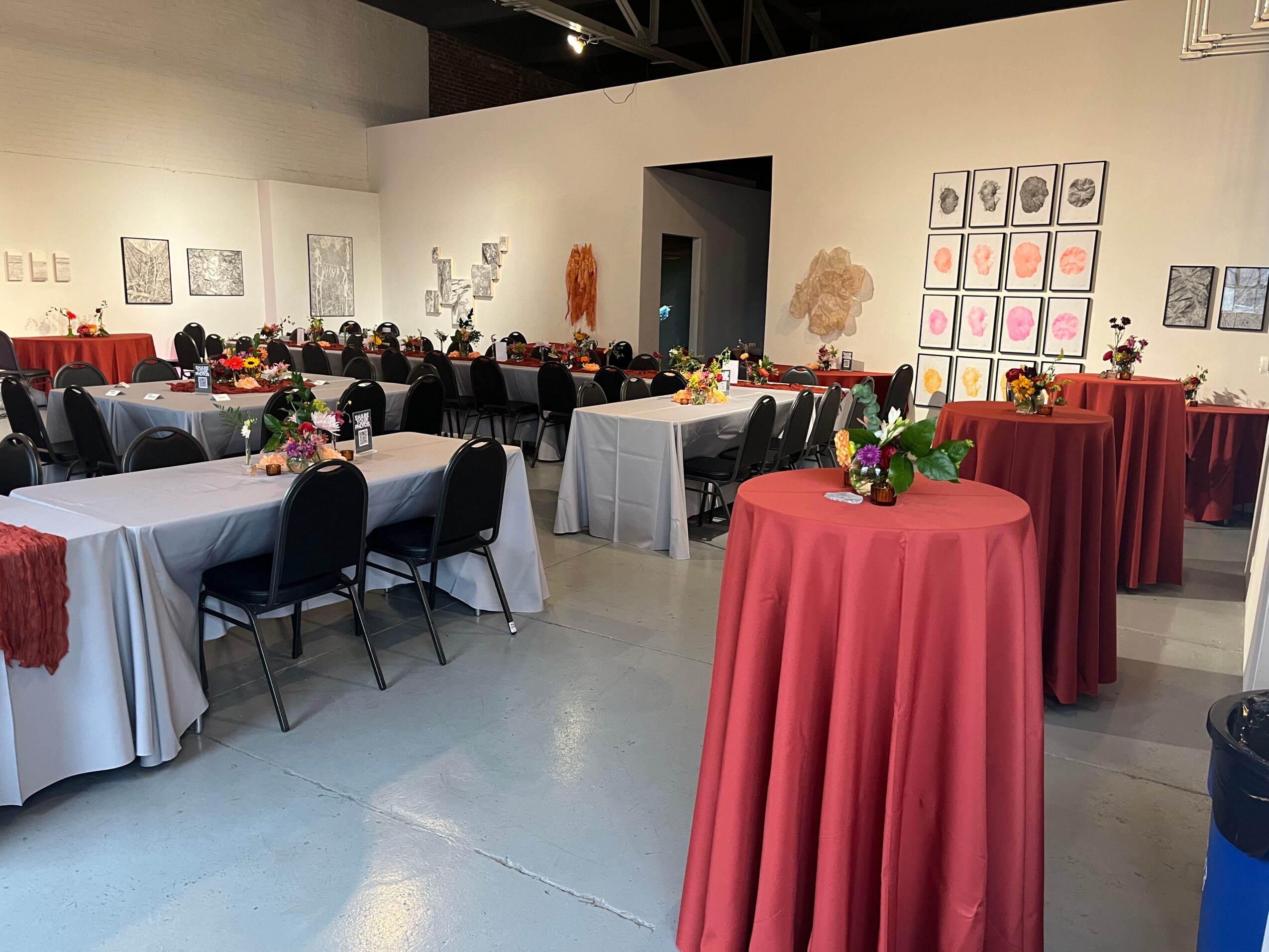 Event space with tables covered in white and red tablecloths, decorated with flowers, and black chairs arranged around them.
