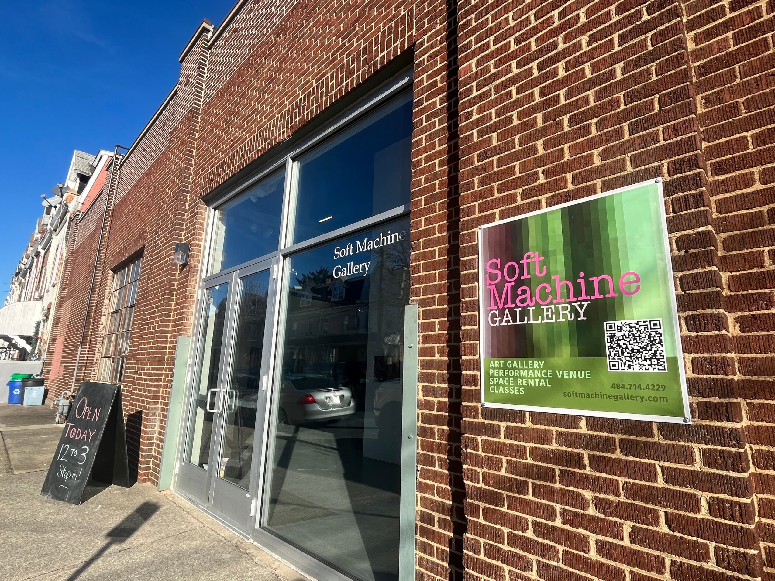 Brick building with glass door and window, sign for Soft Machine Gallery, QR code, and sidewalk.