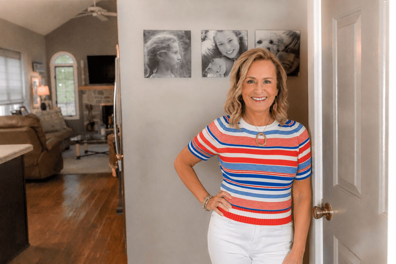 Smiling woman with shoulder-length hair in a striped shirt and white pants standing in a doorway, with a living room in background.
