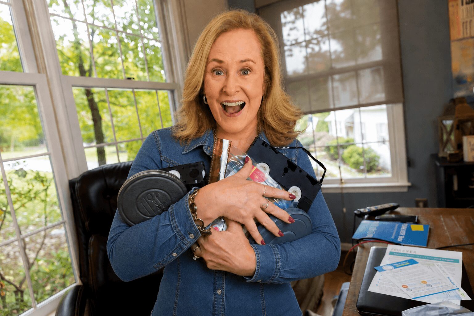 Woman with shoulder-length hair holding a baby, smiling indoors near large windows, with papers and books on a table.