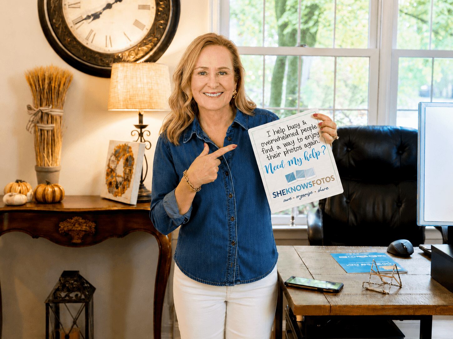 Woman with blonde hair smiling, holding a sign, standing in a room with a clock, lamp, and window behind her.