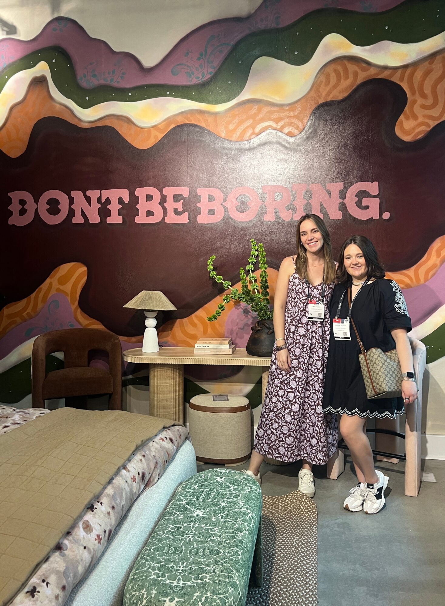Two women stand in front of a colorful wall with the words 'DON'T BE BORING.' They are smiling, with furniture and plants nearby.