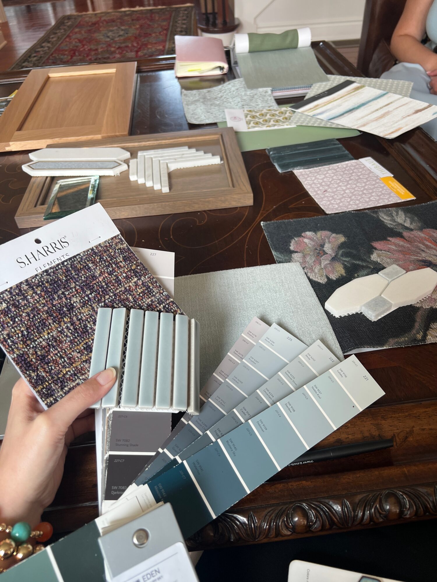 Table with fabric samples, color swatches, and papers, some overlapping, with a person's hand holding fabric swatches in the foreground.
