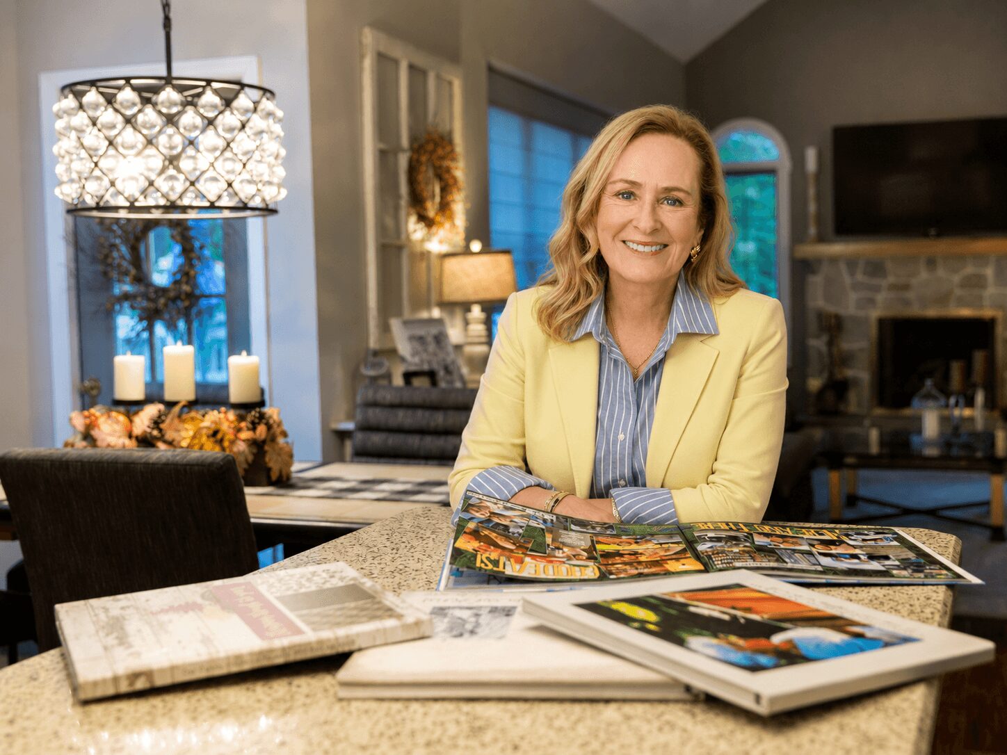 Woman with blonde hair smiling, sitting at a table with magazines in a cozy living room, decorated with candles and artwork.