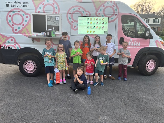 Group of children and adults standing in front of a pink and white mobile classroom van with a screen displaying educational content.