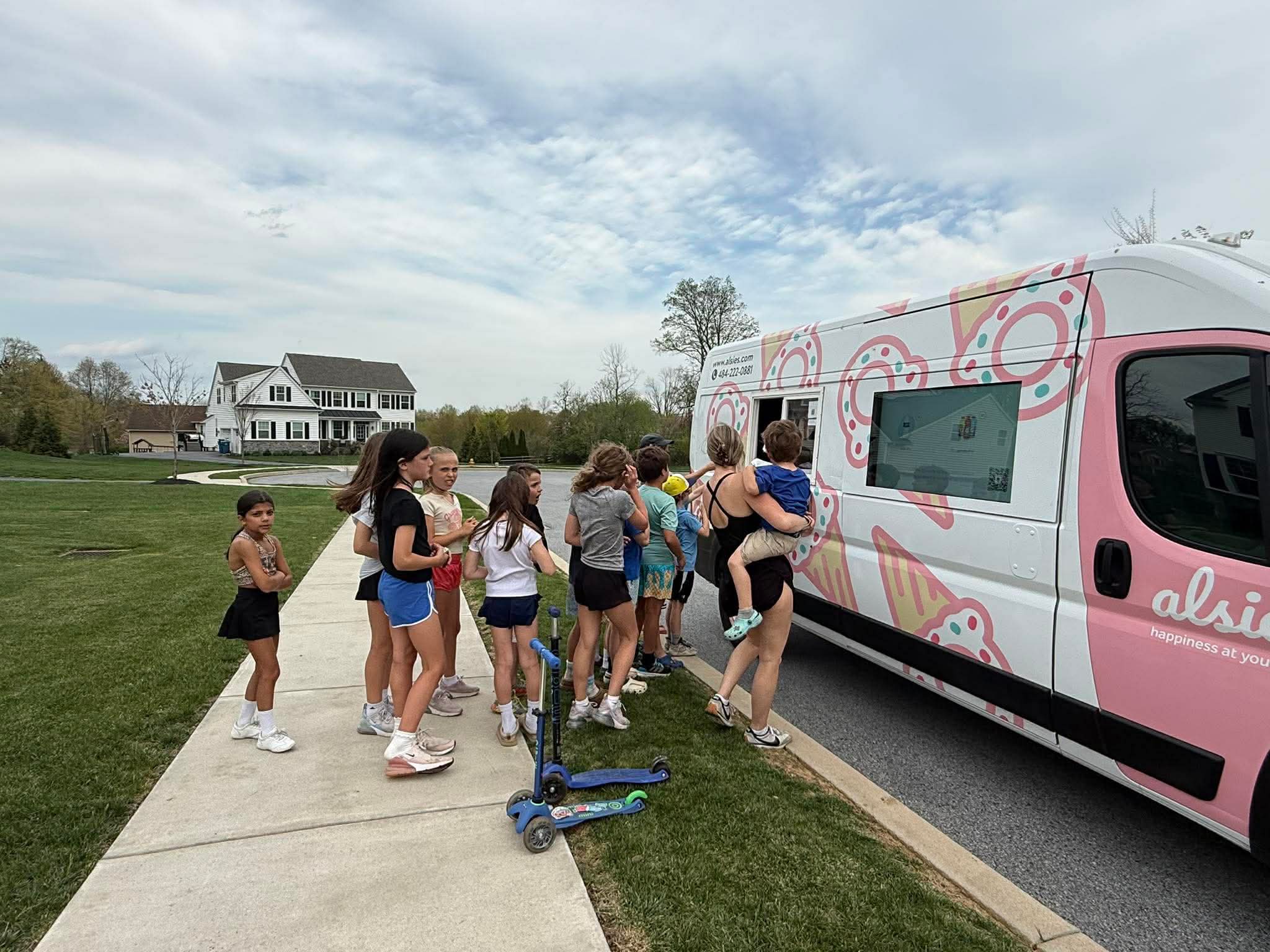Group of children and adults line up to enter a pink and white ice cream truck on a sidewalk in a grassy area, with houses and trees in the background.