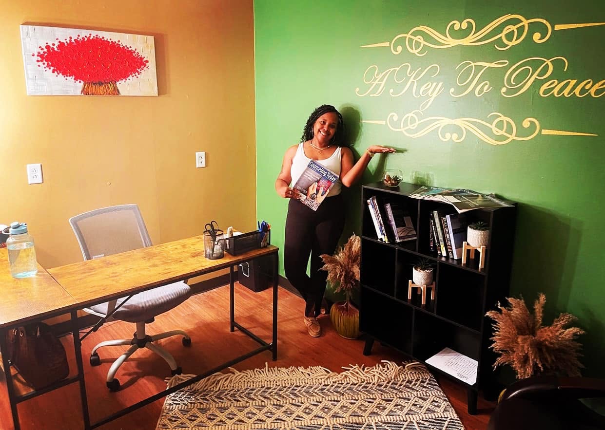 Woman standing in a room with green and yellow walls, holding a book, near a black bookshelf and a table.