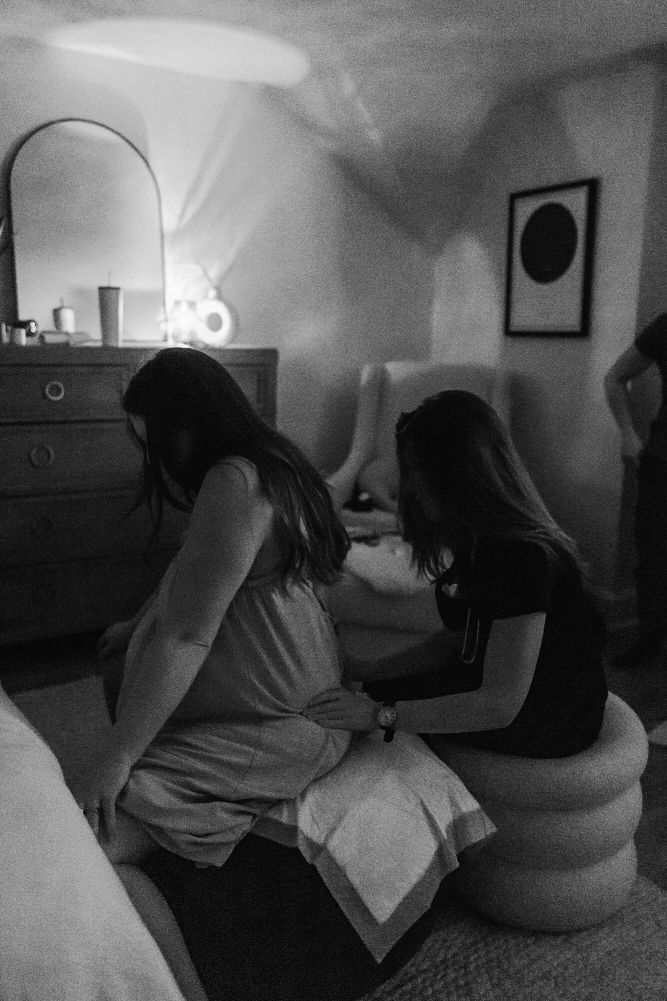 Two women sit on a rounded stool in a dimly lit room, one adjusting the other's clothing, with a dresser and framed artwork in the background.