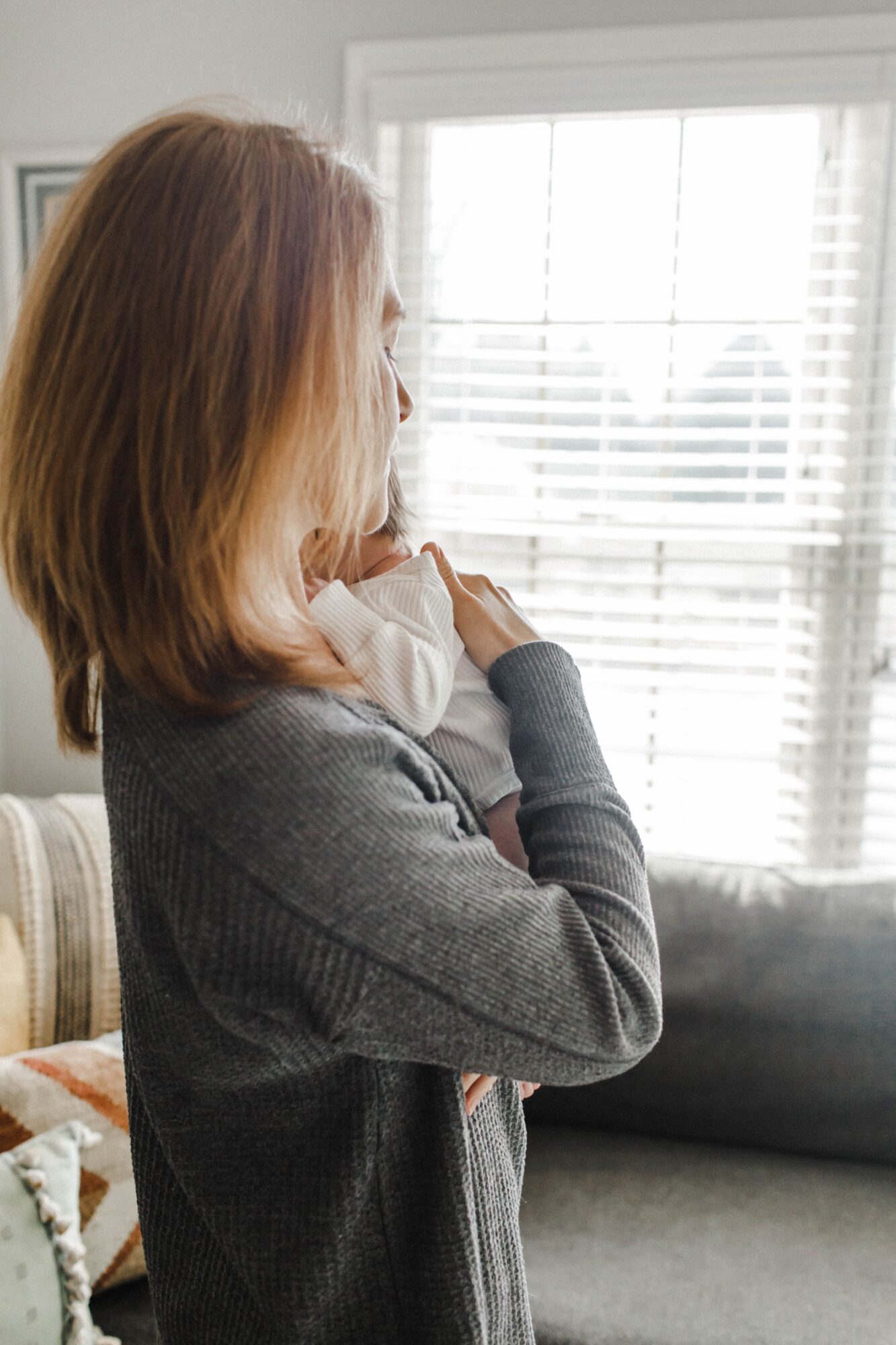 Woman with shoulder-length hair looking out a window with blinds, standing indoors. Natural light illuminates her profile.