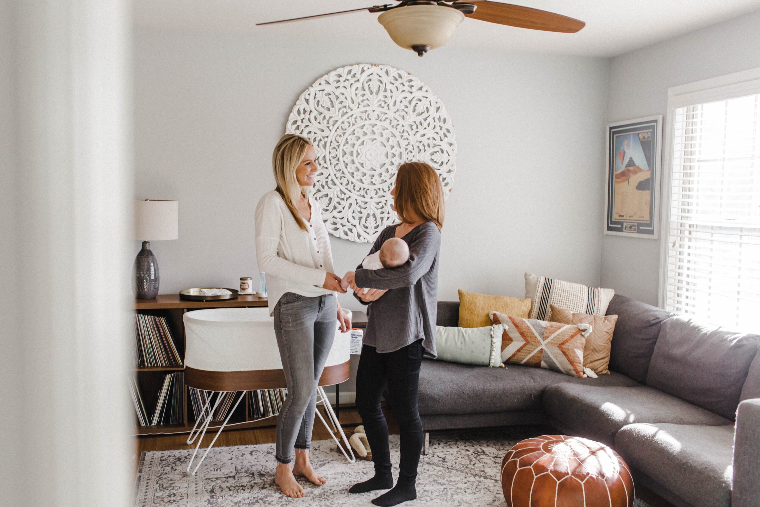 Two women stand in a living room, exchanging a baby doll, with a sofa, pillows, and wall art in the background.