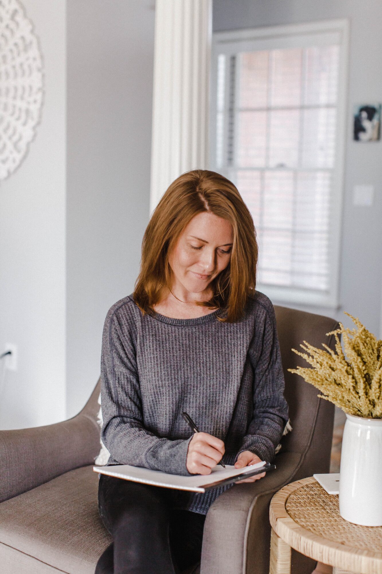 Woman with shoulder-length hair writing in a notebook while sitting on a sofa in a bright room.