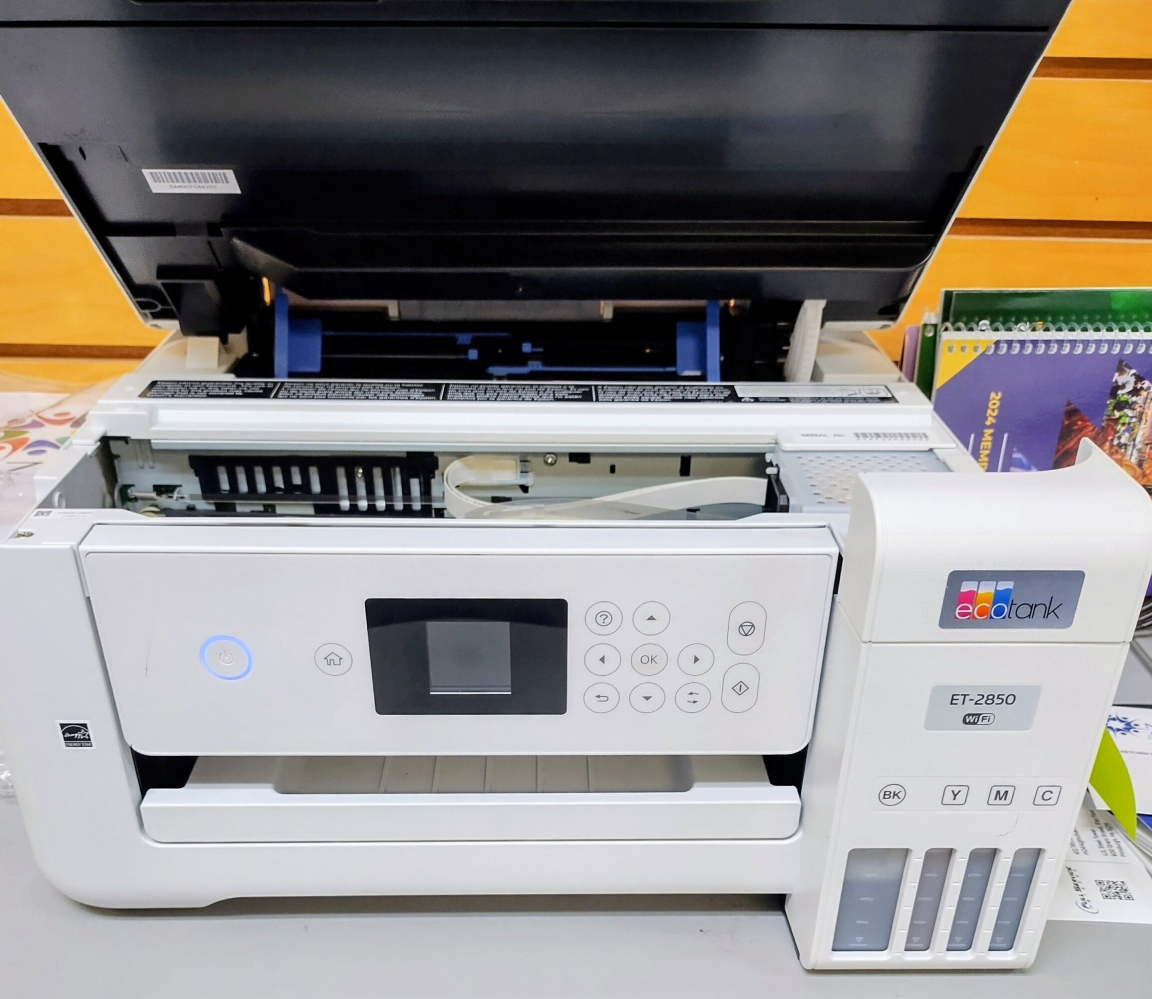 Photograph of a white multifunction printer with an open top, showing internal components, on a table with books nearby.