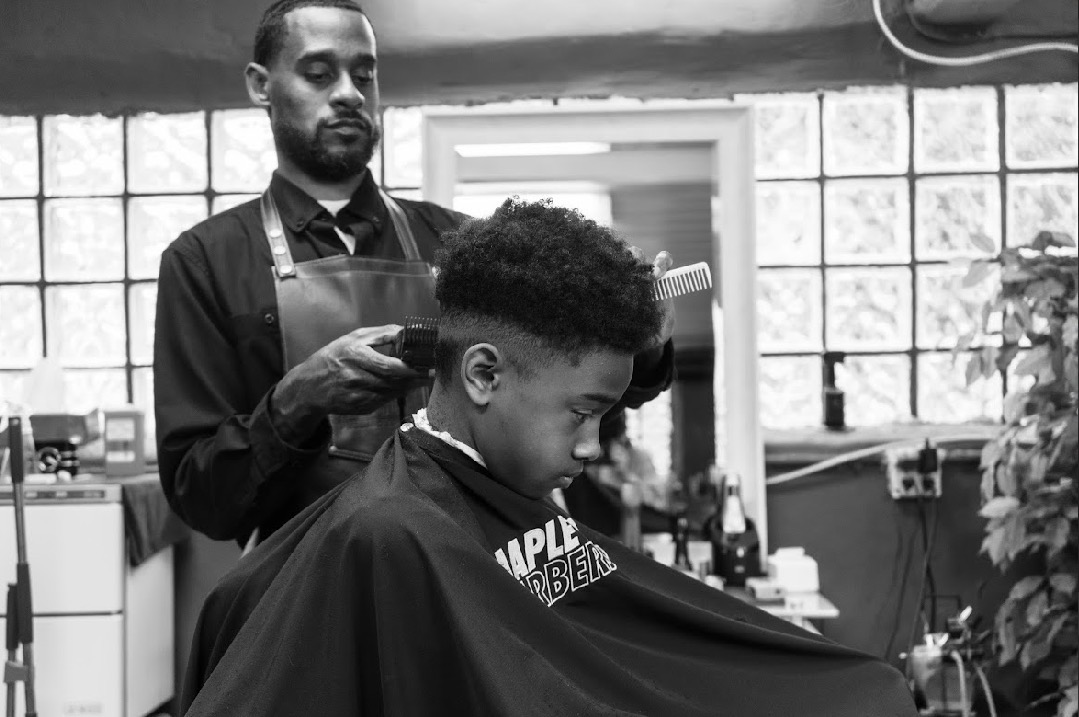 Young person getting a haircut in a barbershop, barber standing behind, black and white photo.