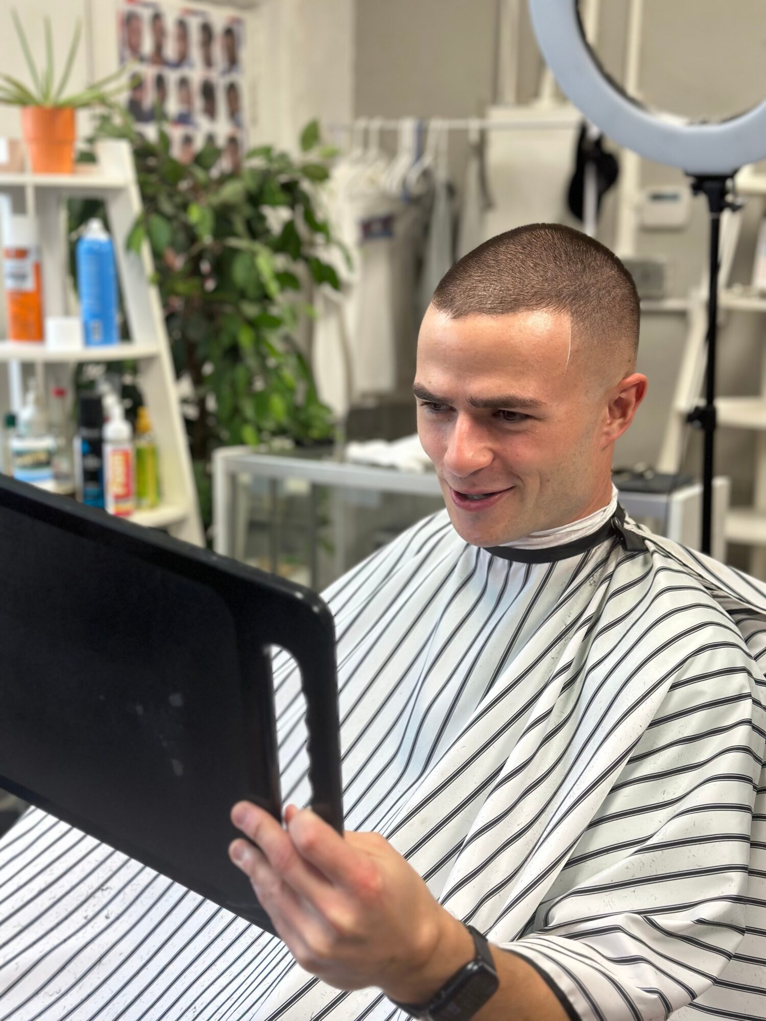 Man with a shaved head wearing a striped barber cape, sitting in a salon, looking at a tablet screen.