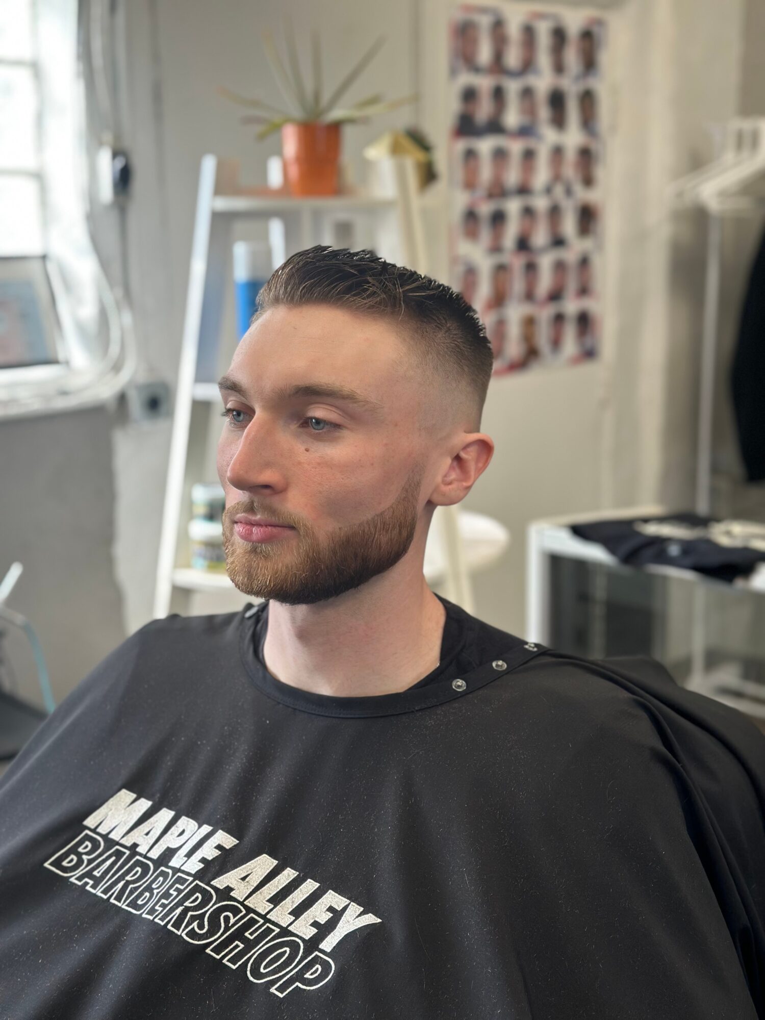 Young man with short hair and beard sitting in barber shop chair, looking to the side, with a poster and plant in background.