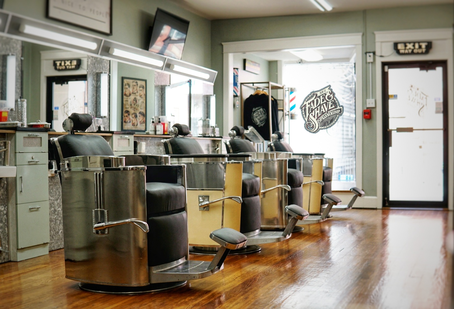 Barber chairs in a salon with mirrors, posters, and a door with a logo outside, wooden floor, and bright lighting.