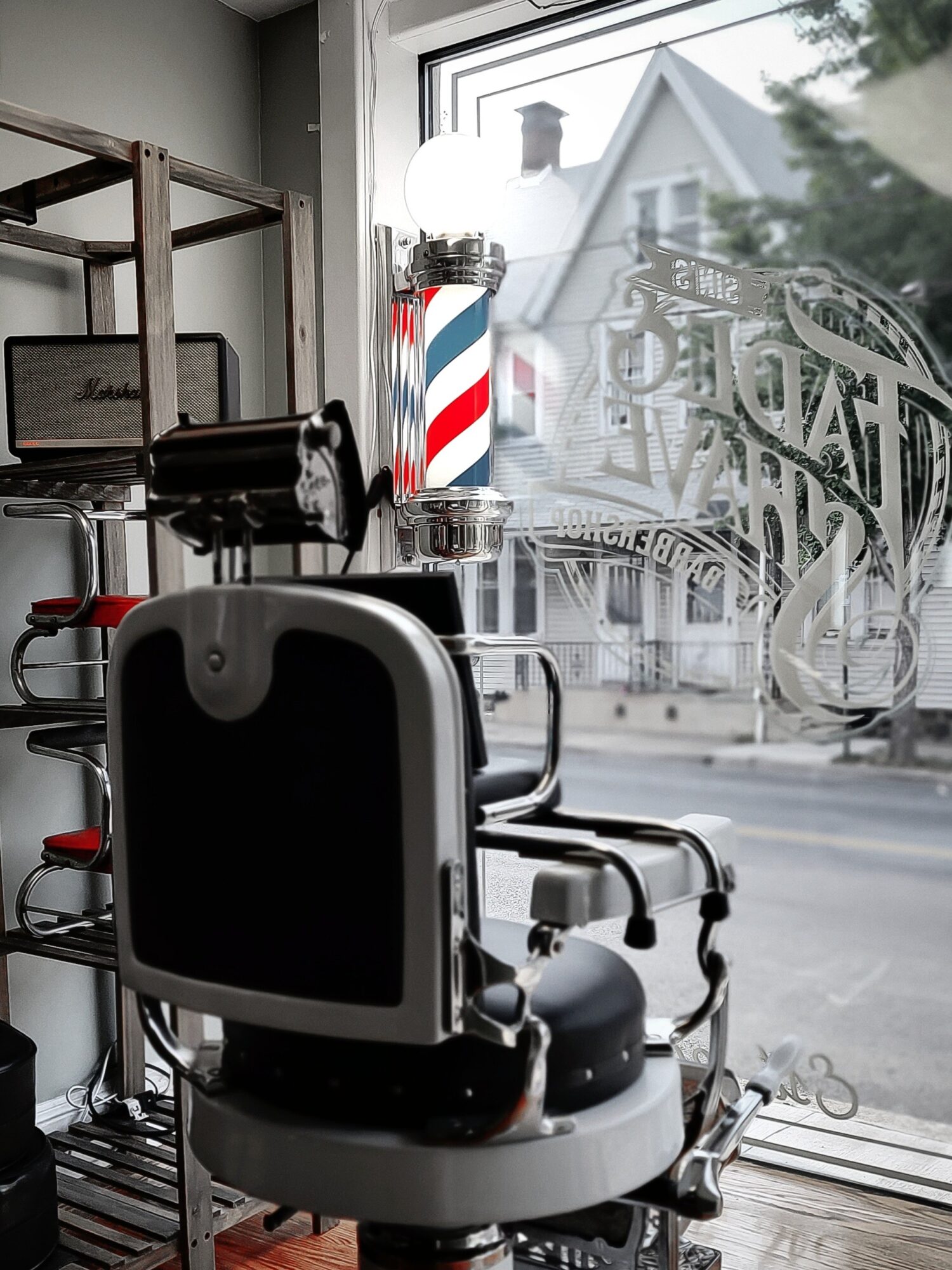 Barber chair facing window with barber tools and a barber pole on the windowsill, outside view of houses and trees.