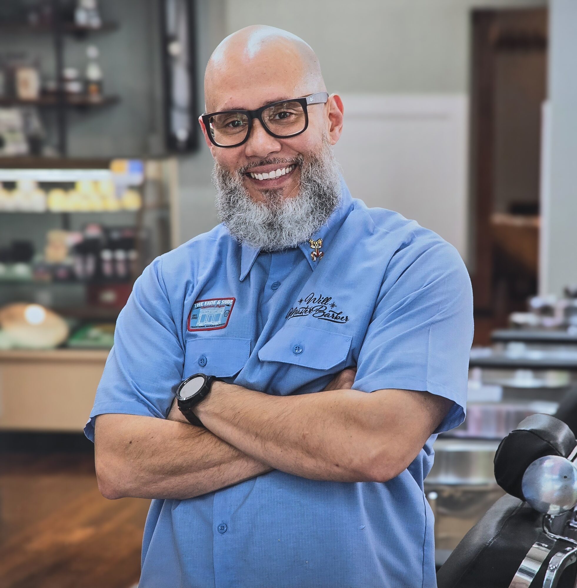 Smiling man with glasses and a beard, wearing a blue uniform, standing with arms crossed in a store or workshop.