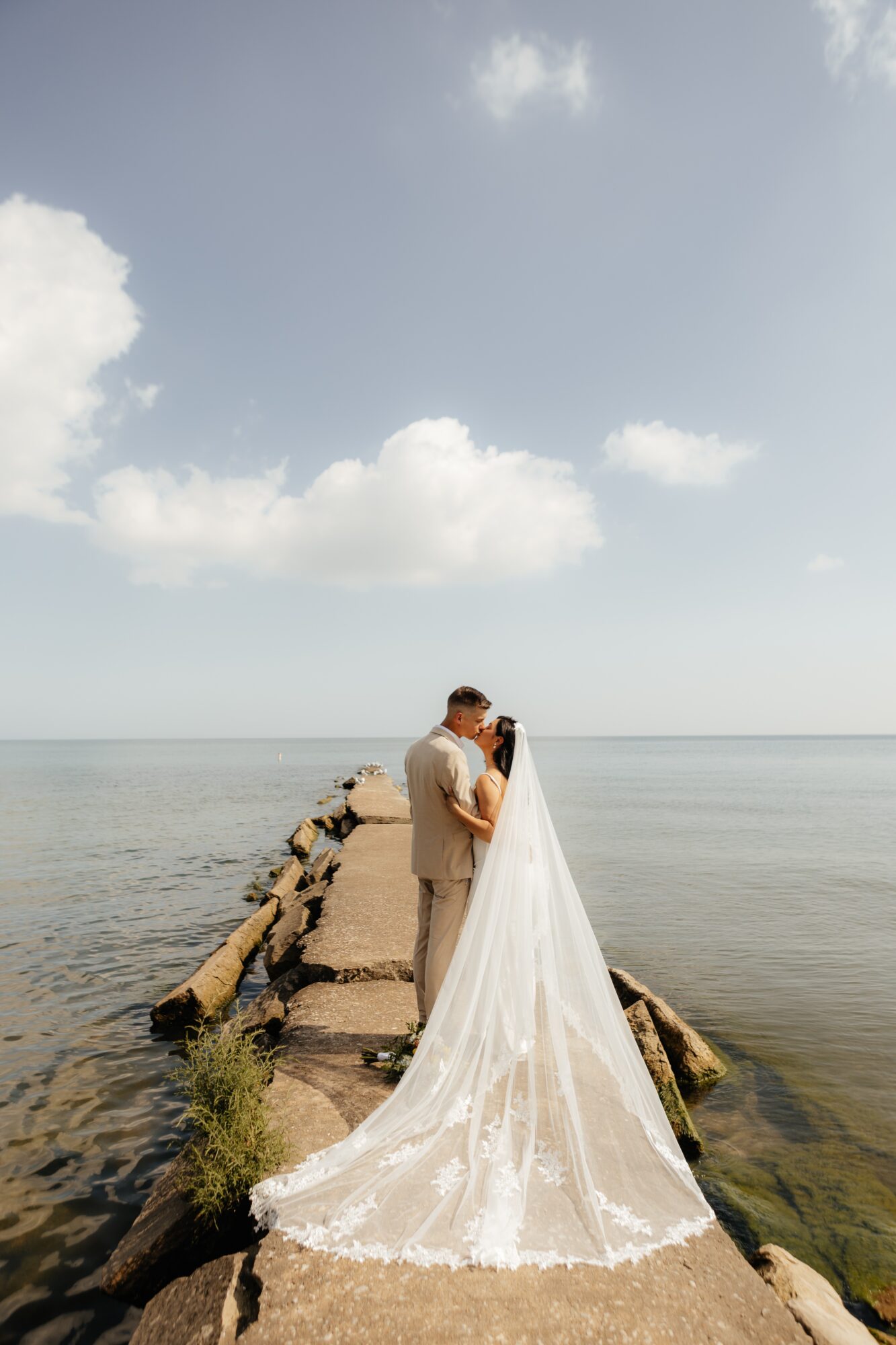 A couple stands on a rocky pier by the water, with a cloudy sky above, embracing each other.