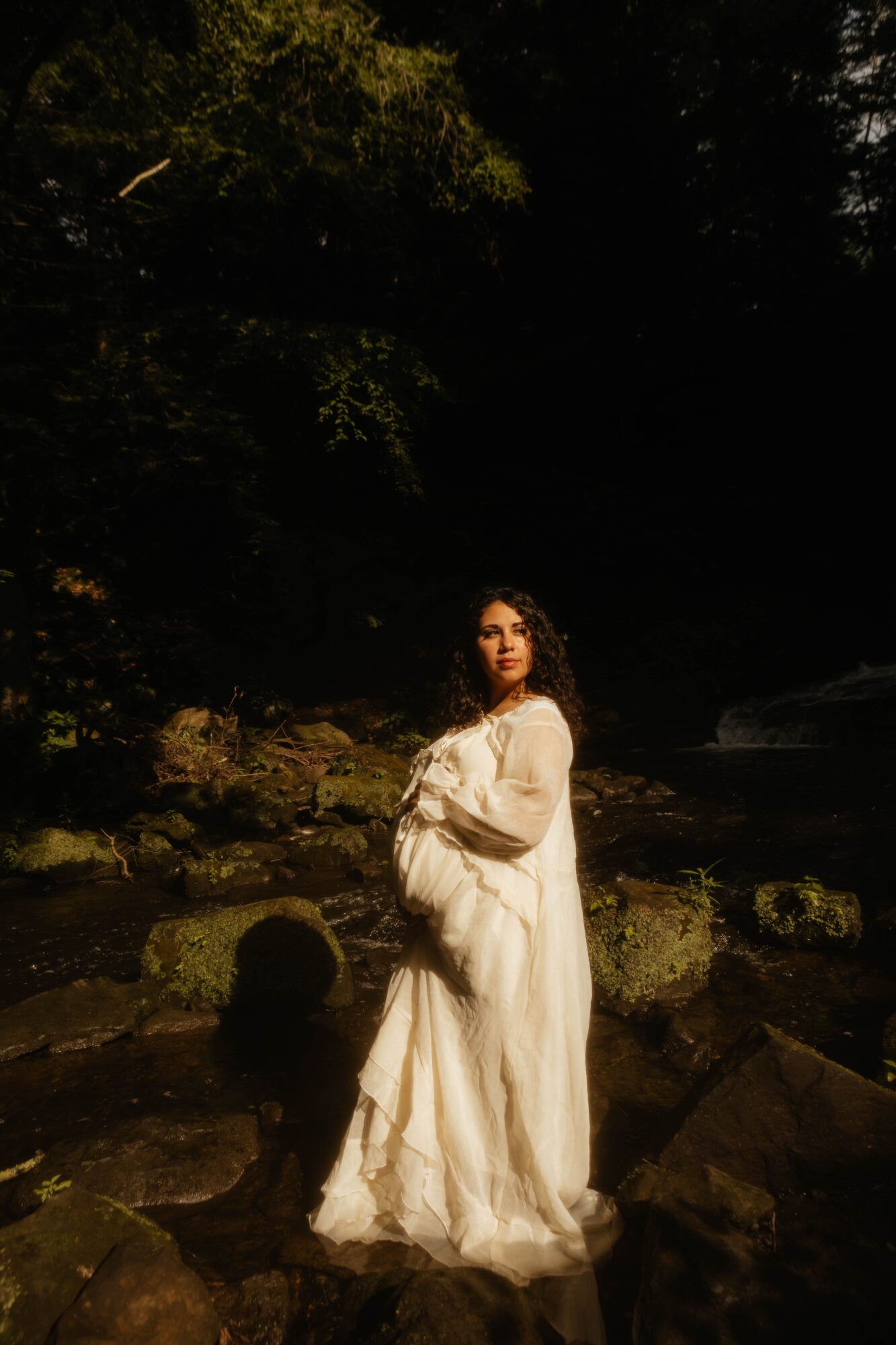 Woman in a long white dress standing outdoors near rocks and water, surrounded by trees and dark foliage.